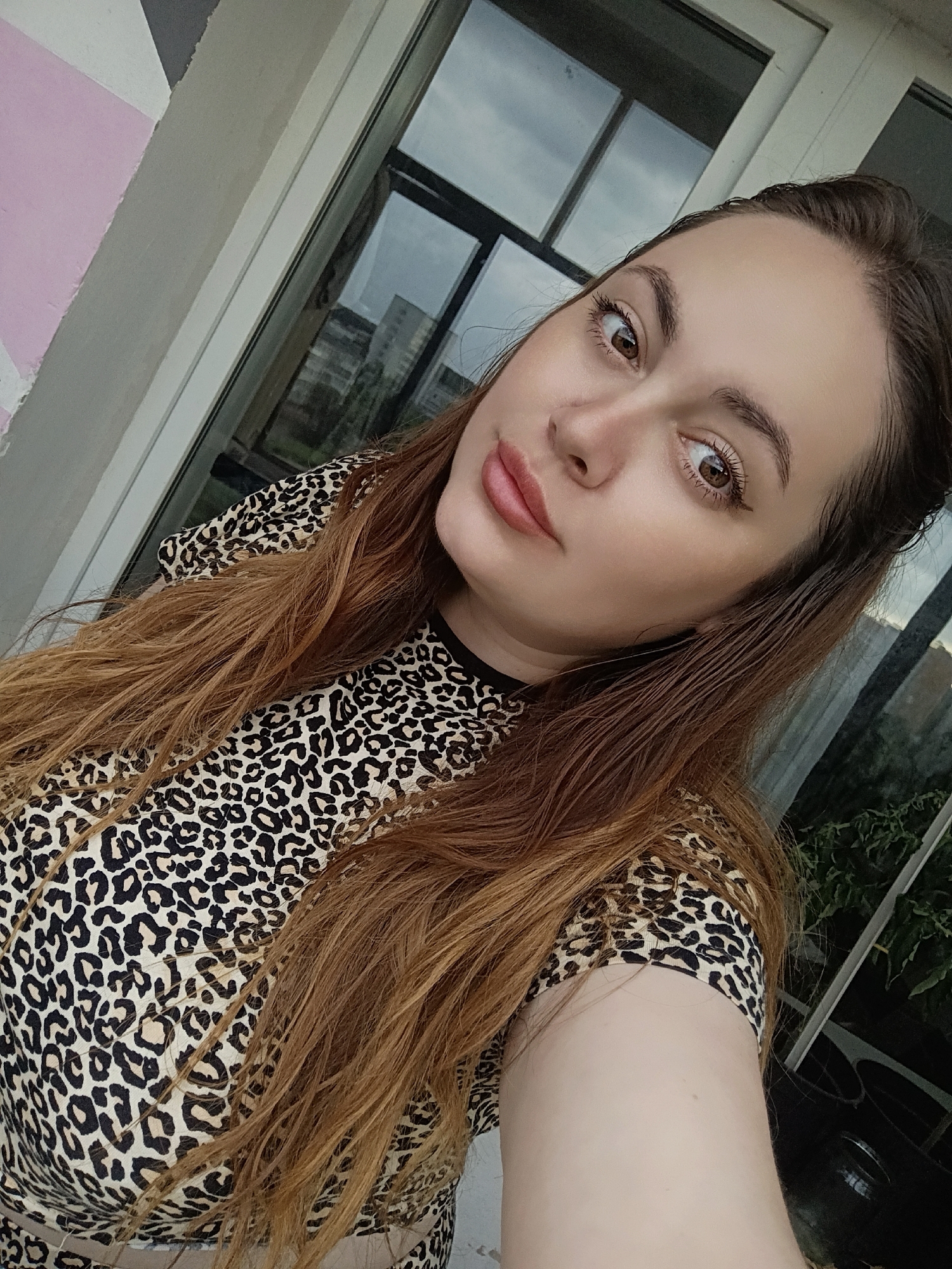 A selfie of a woman is taken at an outdoor location, possibly a balcony, in the city of Chaikovsky, Russia. The woman has long, auburn hair and is wearing a leopard-print top. Her facial features are visible, including her brown eyes, prominent eyebrows, and light pink lips. Behind her, a window with reflections of buildings and sky can be seen. To the left of the woman, a wall with a geometric design in pink and grey is partially visible. The overall lighting suggests it is daytime.