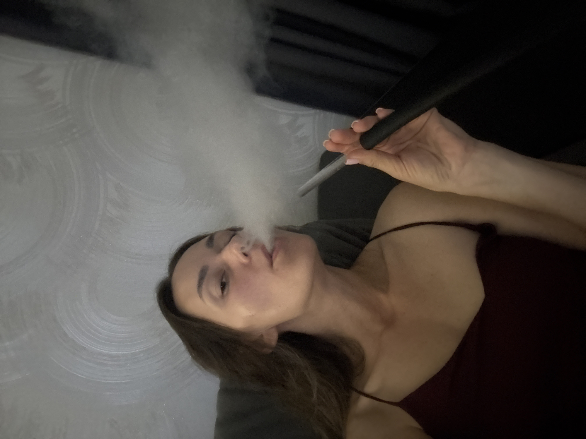A single adult female is depicted indoors in Sochi, Russia, seated or reclining on a dark upholstered surface. She has long, dark brown hair and is wearing a dark red or maroon spaghetti-strap top. Her left hand, with light pink polished nails, holds a black cylindrical mouthpiece or hose, consistent with a hookah or large e-cigarette device. She is actively exhaling a significant volume of thick, white vapor or smoke from her mouth, which partially obscures her face and the background. The background features a light gray wall with a distinctive, brushed, circular texture or pattern. A dark curtain or piece of furniture is visible on the far right of the frame. The lighting appears to be ambient indoor illumination.