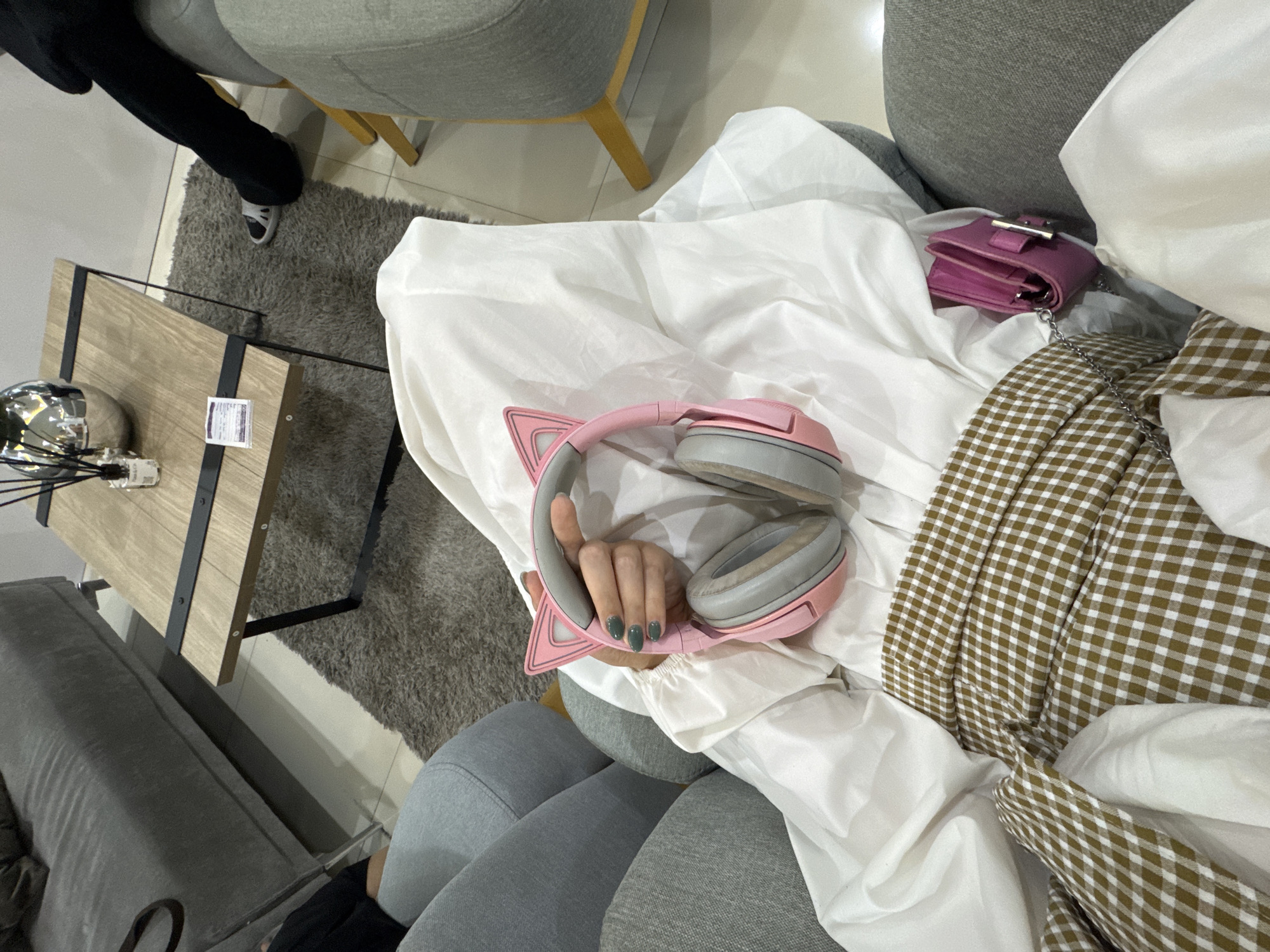 The image shows a close-up view of a person lying down, covered by a white sheet and a brown and white checkered blanket. They are wearing grey leggings and black pants. On top of the sheet, a pair of pink cat-ear headphones with grey earcups rests. A small pink wallet with a silver clasp is also visible. The person's left hand, with dark green nail polish, is resting on the headphones. In the background, a wooden coffee table with black metal accents holds a diffuser with reeds and a silver decorative object. A grey rug is on the floor next to the table, and part of a grey armchair is visible. The setting appears to be indoors, possibly a living room or a lounge area, in a modern style. The lighting suggests it could be daytime. The overall mood is relaxed and casual, with the focus on the personal items laid out on the bed.