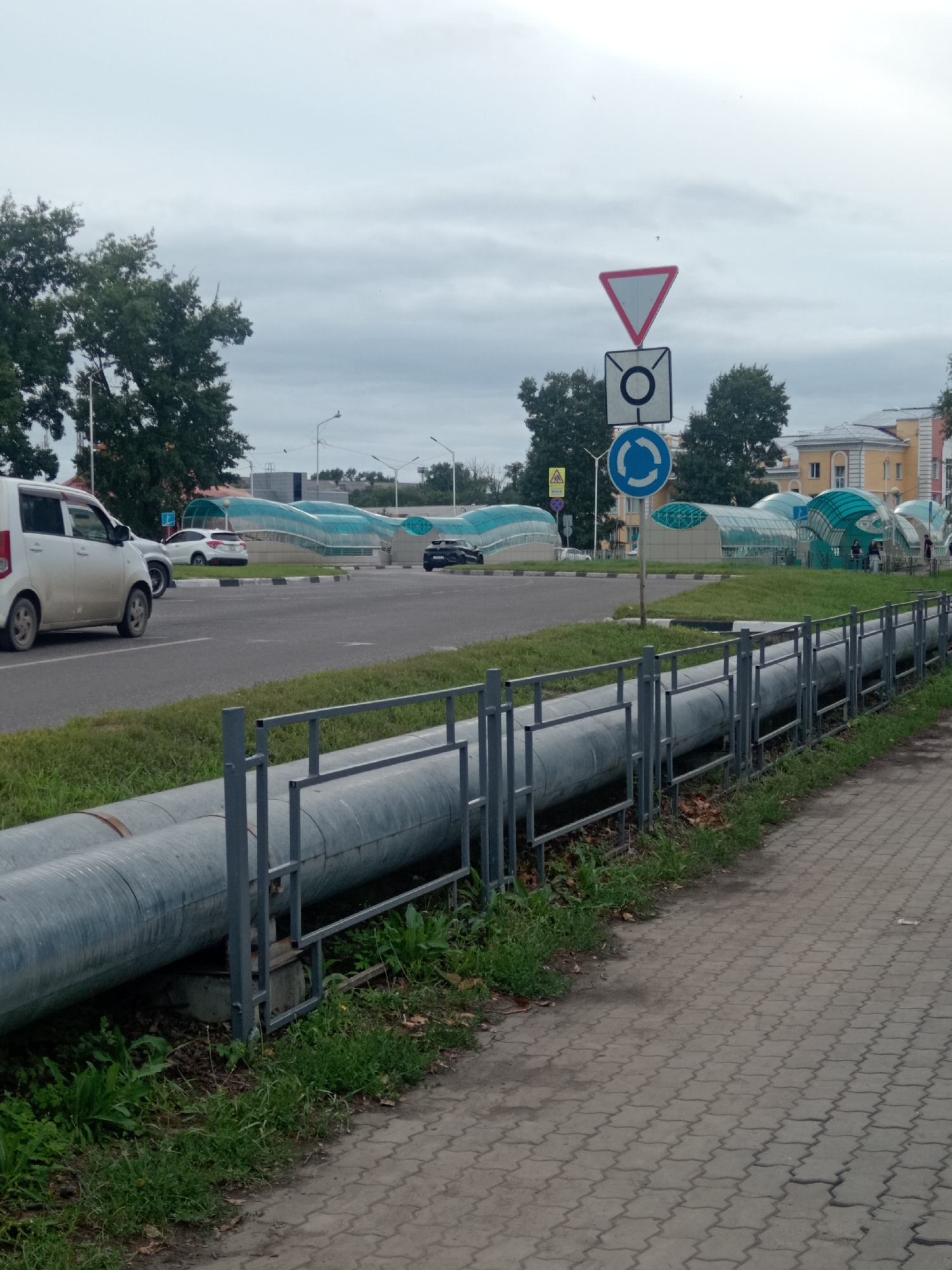 The image depicts an outdoor scene in Belogorsk, Russia, on a cloudy day. In the foreground, a section of a paved walkway is visible, bordered by a metal fence that runs parallel to a grassy area. Behind the fence, two large, parallel industrial pipes are situated on concrete supports. In the middle ground, a road with white lane markings curves to the left. Several vehicles are parked or driving on the road, including a white minivan on the left, a white hatchback further back, and a black car near the center. Beyond the road, a series of modern, tunnel-like structures with green translucent roofs and glass walls are visible, suggesting an urban infrastructure or public space. Traffic signs, including a yield sign and a roundabout sign, are mounted on a pole to the right of these structures. In the background, trees and several buildings with light-colored facades and windows can be seen, including a prominent building with a yellowish facade. The sky is overcast with visible clouds.