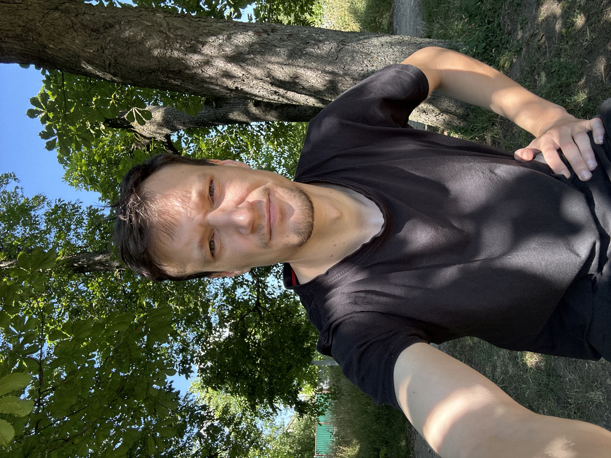 A selfie is taken outdoors, possibly in the city of Makiv, Ukraine, with a man standing near trees. The man has brown hair and is wearing a black t-shirt. His left arm is extended towards the camera, and his right hand is on his hip. The background consists of lush green trees and foliage, with a clear blue sky visible. Sunlight filters through the leaves, casting shadows on the man and the ground. A fence and some dry grass are visible in the distance. The overall setting appears to be a park or a rural area.