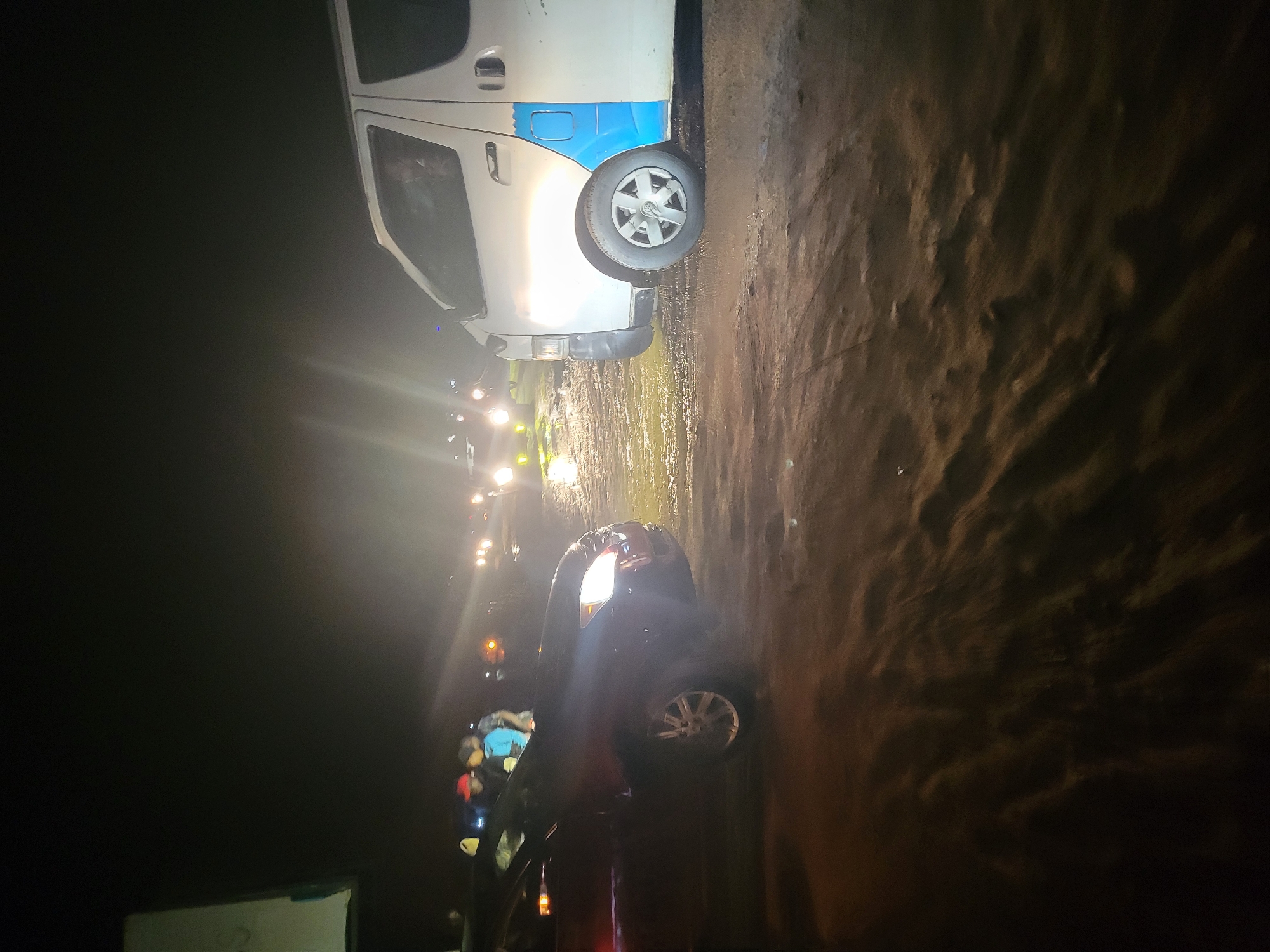 This is a nighttime photograph taken in Kwamba, Nigeria, showing two vehicles on a dirt road. The foreground is dominated by the wet, rutted surface of the road, illuminated by vehicle headlights. To the left is a dark-colored sedan, its headlights casting a bright glow. Several people, some wearing hats, are visible near the sedan. To the right is a white van with blue accents, also illuminated by headlights. The background is dark and appears to be an open area with distant lights suggesting other vehicles or activity. The overall impression is of a scene on an unpaved road at night, possibly a gathering or a traffic stop, with limited visibility due to the darkness and wet conditions.