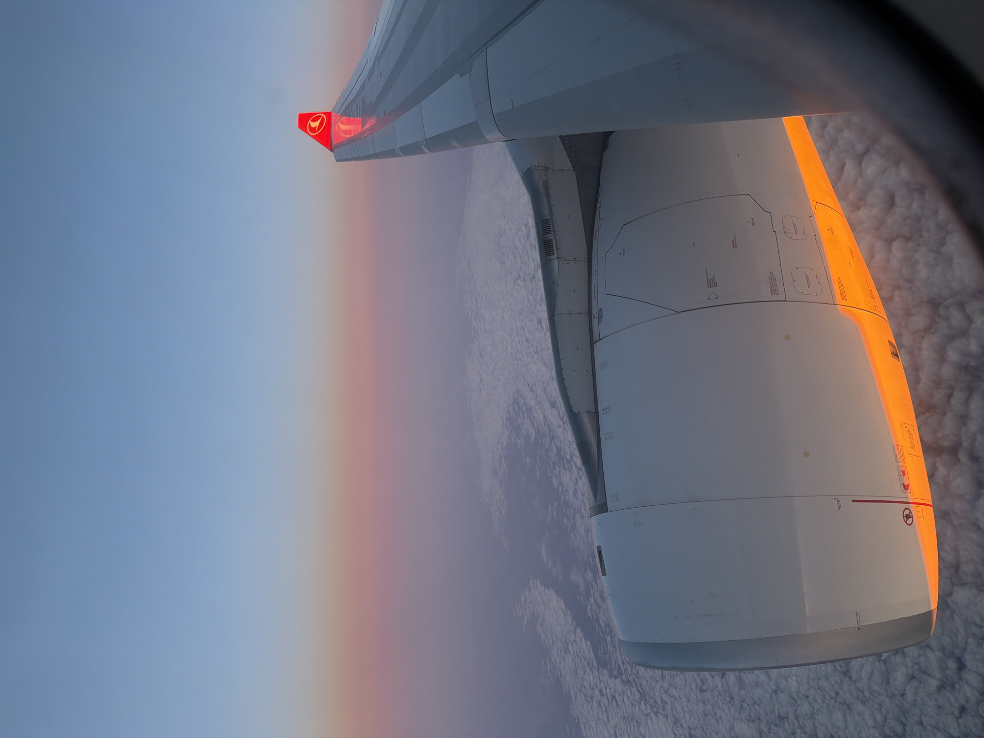 An image taken from the window of an airplane shows the wing and engine against a backdrop of clouds and sky. The aircraft is operated by Turkish Airlines, indicated by the red tail fin with the airline's logo. The sky transitions from blue at the top left to a pinkish hue near the horizon, suggesting sunrise or sunset. The engine is illuminated by the sun, creating a bright orange glow. The scene takes place above the clouds.