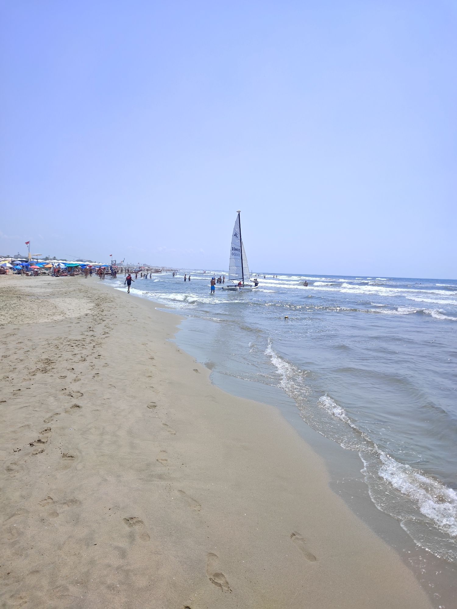 The image depicts a sunny day at a crowded beach in Pietrasanta, Italy. The foreground is dominated by a sandy beach with numerous footprints. Further back, people are seen relaxing under umbrellas and walking along the shoreline. The sea is calm with gentle waves, and a small sailboat with the number "106830" on its sail is visible in the water. The sky is clear and blue. The beach appears to be a popular tourist destination.