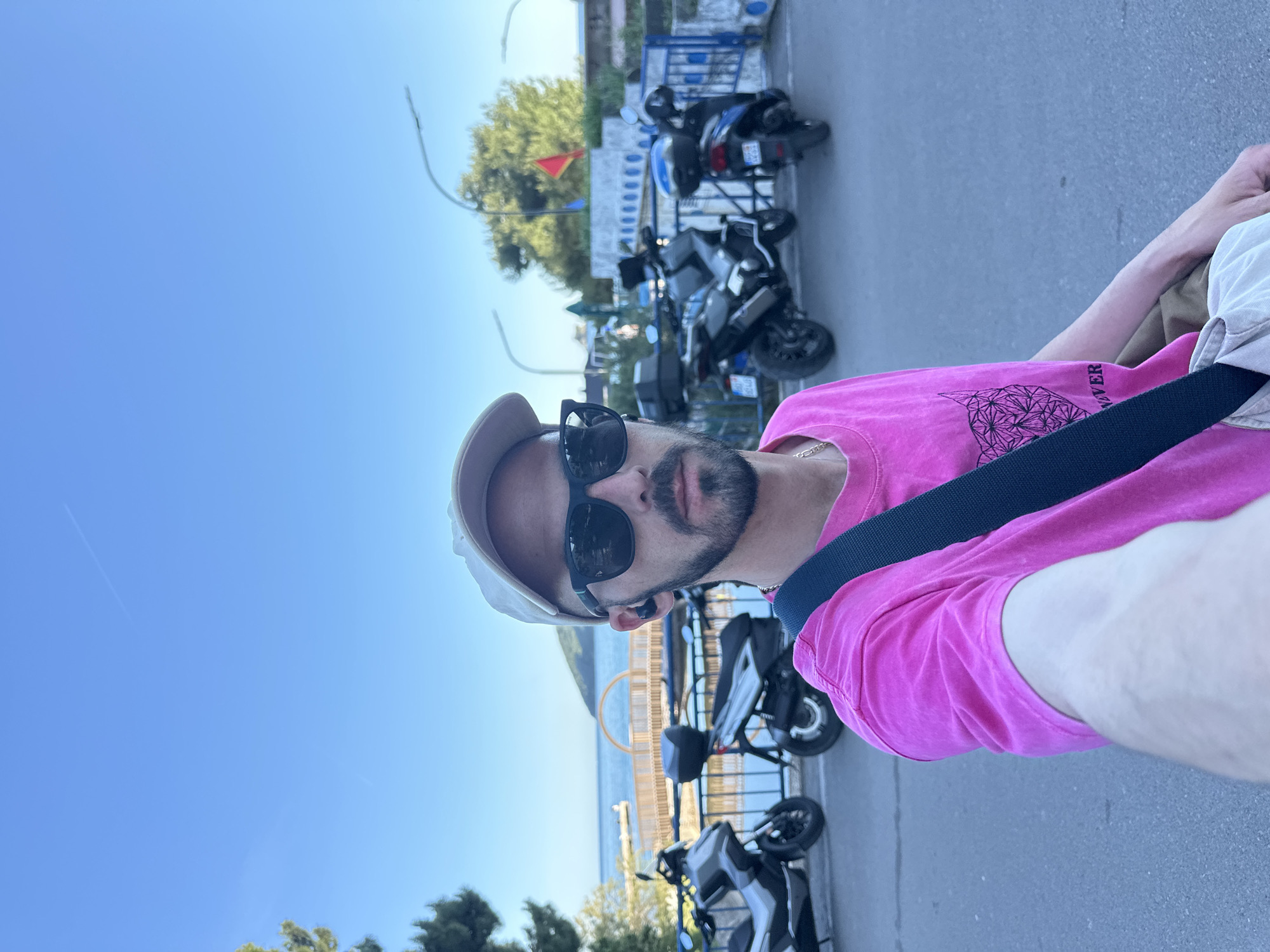 A man is taking a selfie outdoors in Budva, Montenegro. He is wearing a pink t-shirt, beige cap, and sunglasses, with a dark strap across his chest. The background includes a waterfront with the sea, scooters parked along a street, and a blue sky. A blue metal fence is also visible, near a tall sculpture on the water.