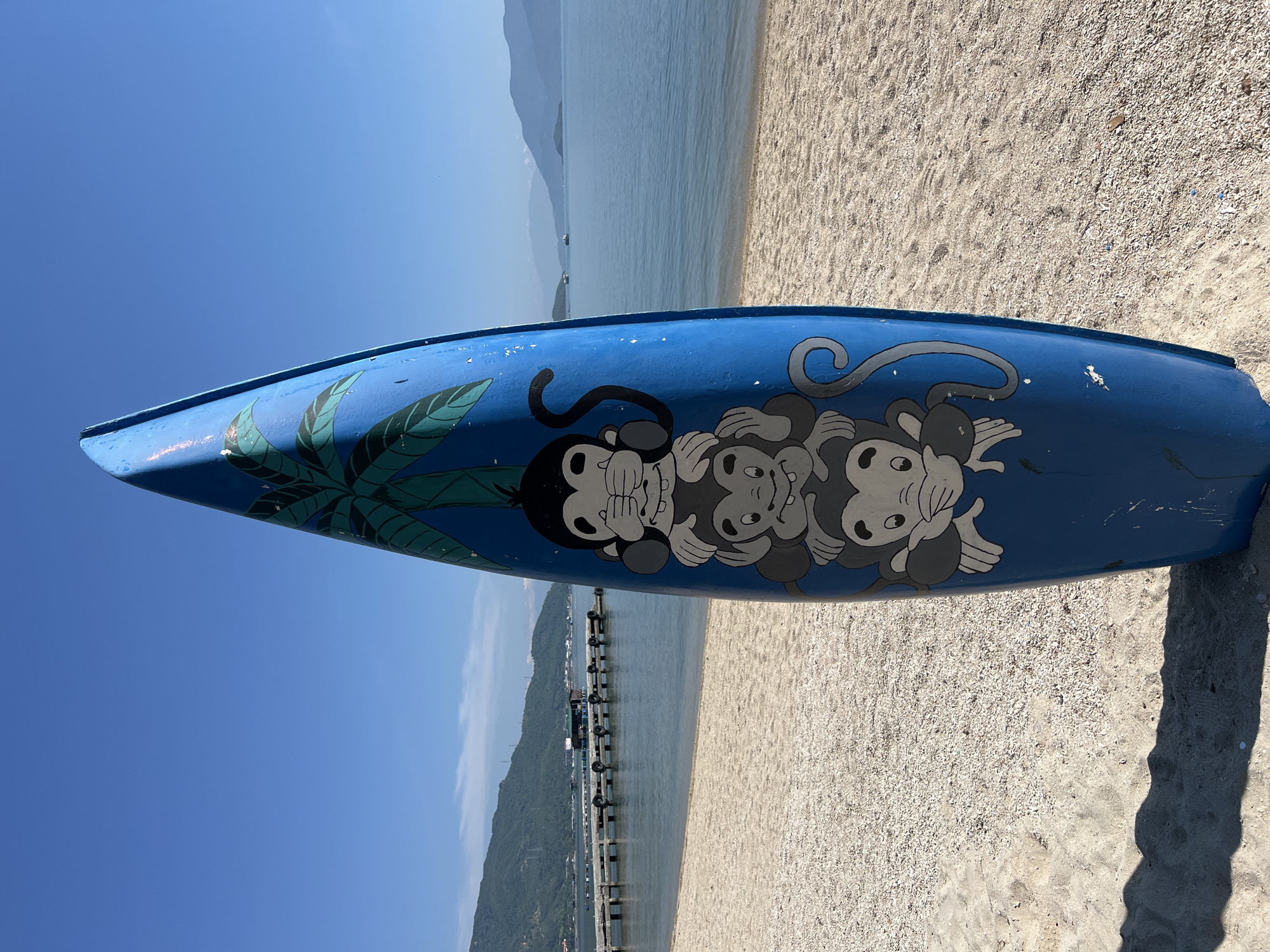 A blue surfboard with a drawing of three monkeys is standing upright on a sandy beach. The monkeys are stacked on top of each other, with the top monkey covering its eyes, the middle monkey covering its ears, and the bottom monkey covering its mouth. A palm tree is drawn behind the monkeys. The beach is made of sand and small pebbles. In the background, there is a calm body of water, a pier with structures on it, and lush green hills. The sky is clear and blue. The location is the city of Bac Nha Trang Ward, Vietnam.