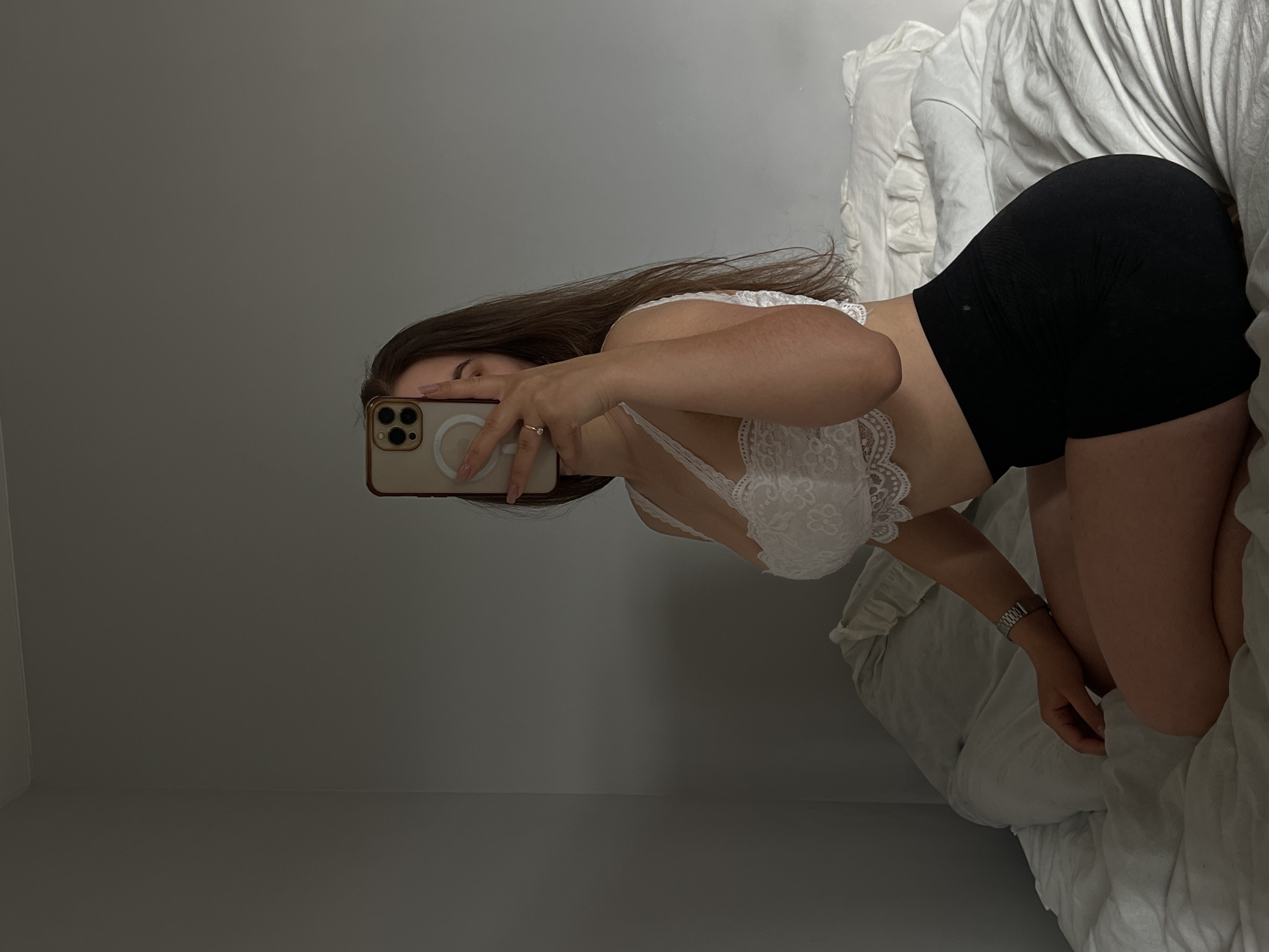 A young woman is sitting on a bed, taking a selfie in a mirror. She is wearing a white lace bralette and black shorts. The room is dimly lit, with a grey wall in the background and white pillows and sheets on the bed. The woman's face is obscured by her phone, but her hand is visible, wearing a ring. She has long brown hair. The overall impression is one of casual intimacy. There is no indication of the location being Ternopil, Ukraine.