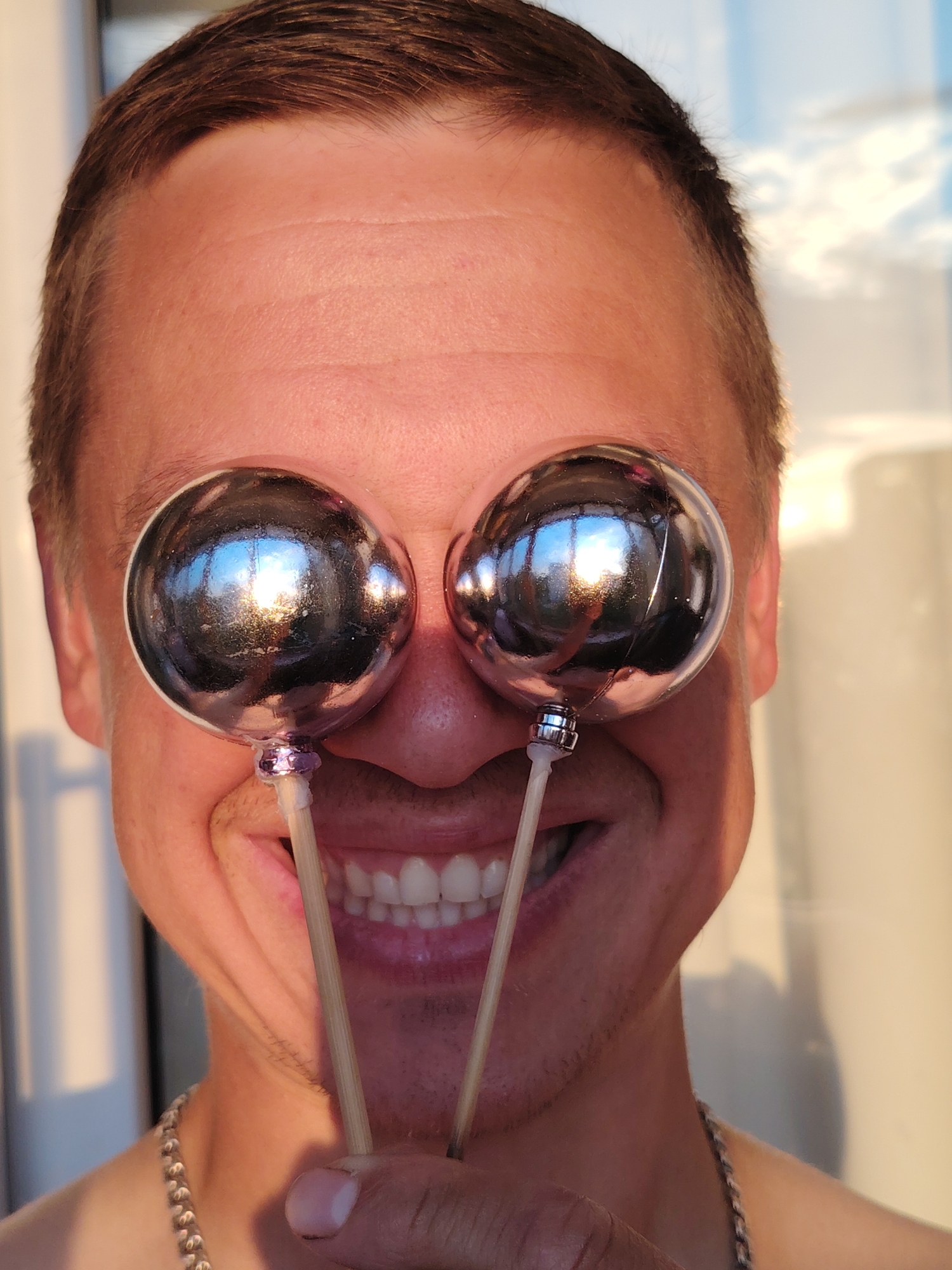 The image is a close-up of a man's face, from the nose up. He is smiling broadly and holding two small, shiny, spherical objects on sticks in front of his eyes. The objects are reflective and show the sky, with clouds, and a bright light source. The man has short brown hair and is wearing a chain around his neck. The background appears to be indoors, near a window, with soft, warm lighting. There is no indication of Chaikovsky, Russia, in the image.