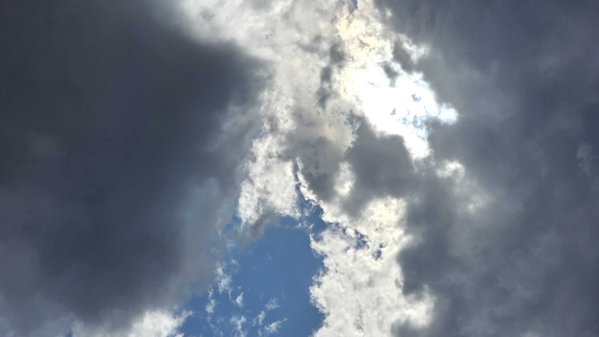 A dramatic sky over Kyiv, Ukraine is filled with a mix of dark, ominous storm clouds and patches of bright, sunlit sky. The clouds are dense and textured, with shades of grey and deep charcoal dominating the upper portion of the image. In contrast, a bright, almost blinding light shines through a gap in the clouds towards the center-right of the frame, casting ethereal rays. A small sliver of clear blue sky is visible in the upper left quadrant, hinting at a break in the weather. The overall impression is one of a dynamic and potentially turbulent sky, common during transitional weather patterns. No people, structures, or specific landmarks are visible, as the image is focused solely on the atmospheric conditions.