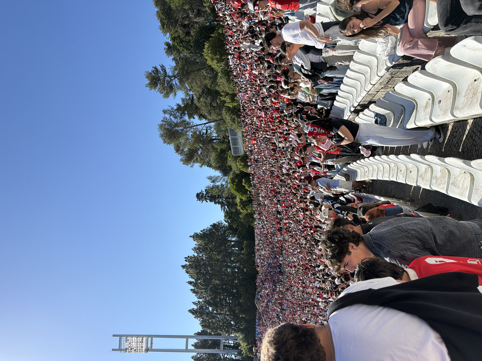 The image shows a large crowd of people densely packed in a stadium setting. The majority of the crowd is dressed in red and white clothing, with some individuals wearing hats or scarves of the same colors. The location is the Estádio Nacional in Oeiras, Portugal, indicated by the presence of the stadium lights and the surrounding trees. The rows of white stadium seats are visible. The scene is set outdoors on a clear day with a bright blue sky.