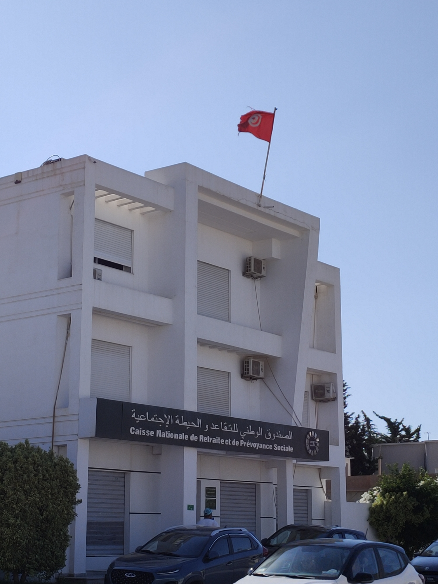 The image shows the front of the Caisse Nationale de Retraite et de Prévoyance Sociale building in an urban setting. The building is white, with multiple floors and several windows that are covered with white shutters. There are also air conditioning units visible on the exterior walls. A flag of Tunisia, red with a white circle containing a red crescent and star, flies from a flagpole on the roof of the building. Below the flag, on the facade of the building, is a sign with Arabic and French text that reads "الصندوق الوطني للتقاعد و الحيطة الاجتماعية Caisse Nationale de Retraite et de Prévoyance Sociale". Several cars are parked in front of the building, including a dark-colored SUV and a white hatchback. A large green bush is visible on the left side of the image. The sky is clear and blue, suggesting it is daytime.