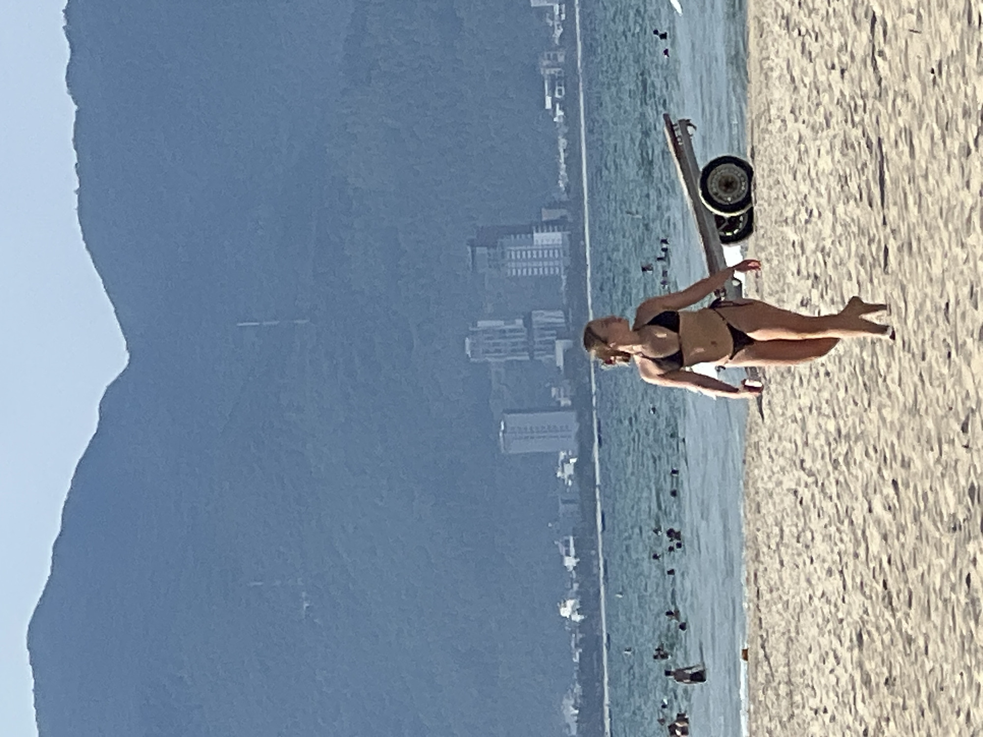A young woman in a black bikini walks on a sandy beach towards the right of the frame, with the ocean and a city skyline in the background. She has blonde hair pulled back and is facing left in profile. In the water behind her, many people are swimming or wading. To her right and slightly behind her, a trailer with two wheels rests on the sand, partially submerged in the water. The city skyline features several tall buildings, including two prominent high-rises. In the far background, a large, blue-grey mountain range dominates the horizon. The scene is consistent with a view of the coastline of Da Nang, Vietnam, likely from one of its popular beaches such as My Khe Beach. The lighting suggests it is a sunny day.