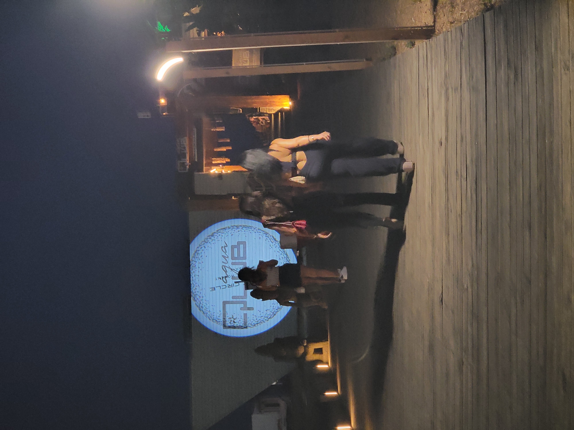In Quarteira, Portugal, a nighttime scene unfolds on a wooden boardwalk leading towards a projected circular logo displaying "CIRCLE of aqua CLUB." Two women, viewed from behind, are walking away from the camera on the boardwalk. The woman on the left has dark, long hair and is wearing a dark top and dark pants. The woman on the right has darker, curly hair and is wearing a dark tank top and dark pants. In the distance, another woman with dark hair, wearing a checkered top and dark shorts, is walking towards the projected logo. Several outdoor lights are visible along the left side of the boardwalk, casting light onto the wooden planks. The background consists of a dark, dusky sky and the facade of a building with the projected logo.