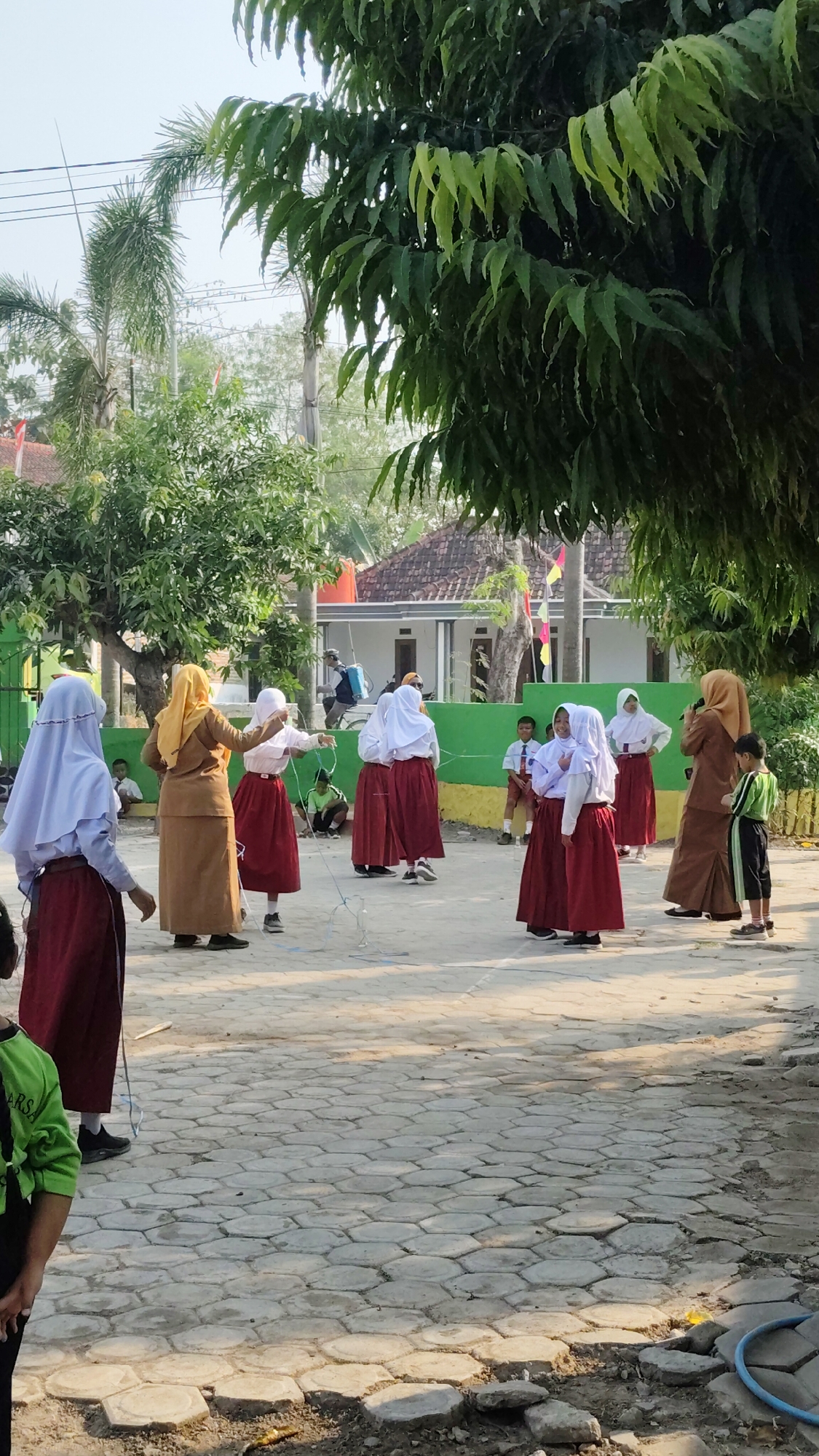 This image depicts an outdoor scene at what appears to be a school in Banjarsari, Indonesia. Several young girls, dressed in school uniforms consisting of white hijabs, white tops, and maroon skirts, are engaged in a group activity. One girl in the foreground is wearing a light purple hijab and a maroon skirt. To her right, a teacher or adult woman, wearing a brown hijab and a long brown skirt, is interacting with a student. Another student in a white hijab and maroon skirt is holding a rope, possibly for a game. In the background, more students in similar uniforms are visible, along with a person on a bicycle with a blue tank on their back, who might be a staff member or a delivery person. The environment includes a paved courtyard with hexagonal and rectangular stone patterns, a green fence, trees, and buildings in the distance. The lighting suggests it is daytime.