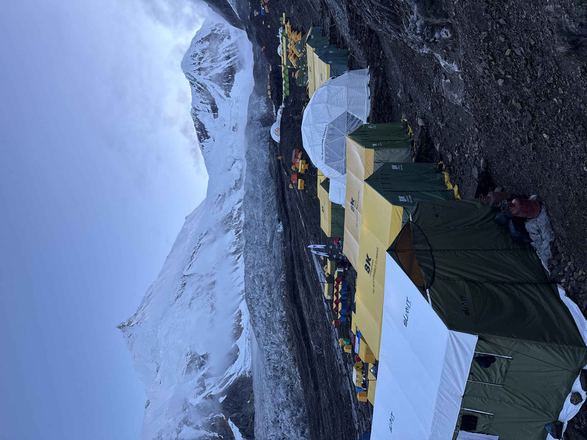The image shows a mountain campsite at an early hour, with tents spread across a rocky, dark-colored terrain. In the background, a vast, snow-covered mountain range dominates the scene, with some clouds obscuring the upper peaks. A glacier is visible on the lower slopes of the mountains. Numerous tents of various sizes and colors are pitched, including large white and yellow structures, as well as smaller multi-colored tents. Some of the tents have logos, such as "SUMMIT" and "8K EXPEDITIONS THE RESPONSIBLE CLIMBERS." One large white dome tent is also present. Two red gas cylinders are visible next to a green tent in the foreground. The overall impression is of a base camp for a high-altitude expedition, likely in a mountainous region. The location is specified as Samagaon, Nepal.