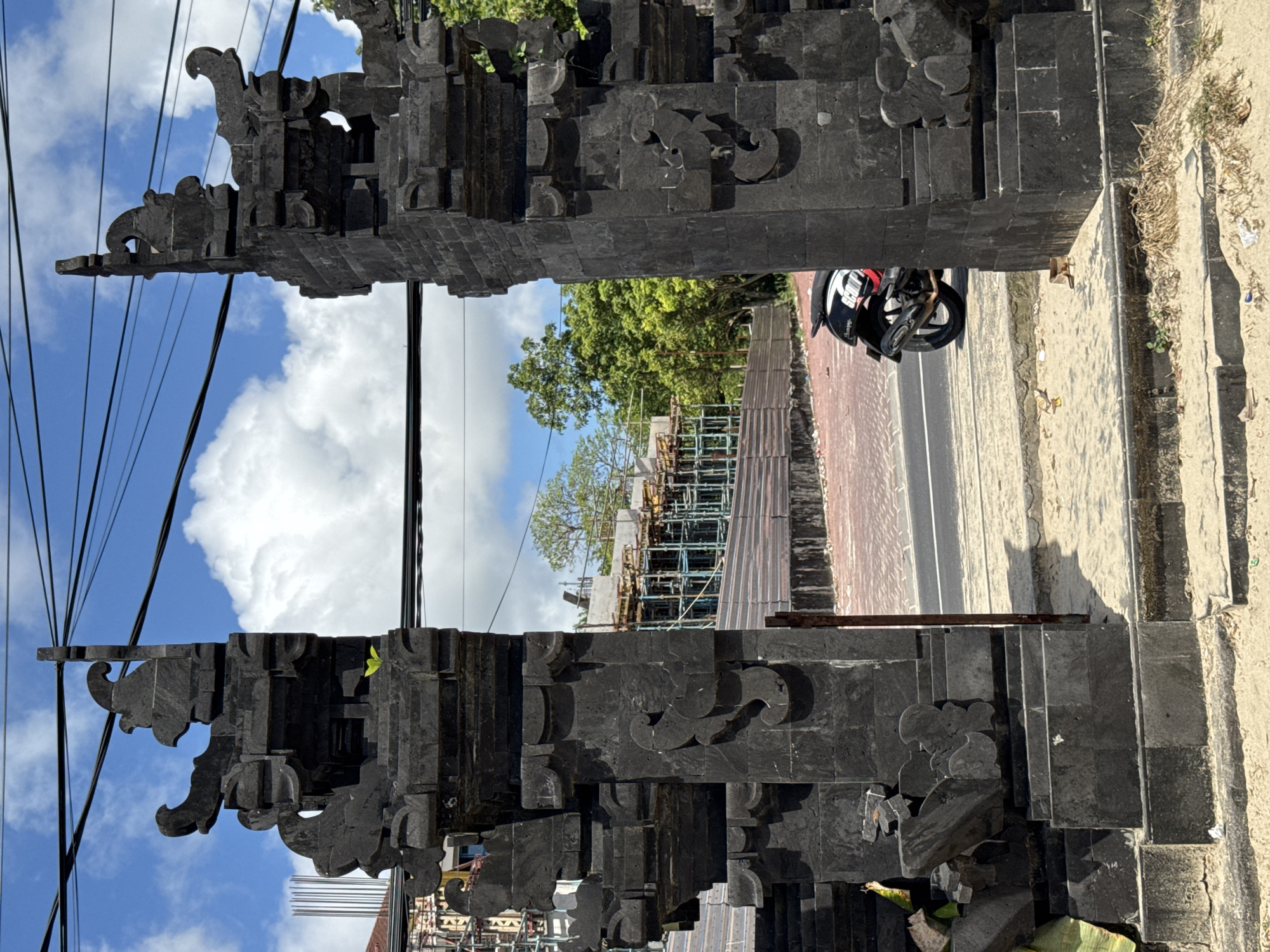 Captured in the urban setting of Kuta Selatan, Indonesia, the image features a structure constructed of dark stone blocks, possibly a gateway or decorative wall. The stone pieces are meticulously carved with intricate designs and arranged in a layered formation. A motorcycle with a helmet attached is parked on the road, next to this stone structure. Utility wires stretch across the sky, which displays a mix of clear blue and fluffy white clouds. In the background, there is scaffolding, suggesting ongoing construction work. The photo is taken at street level, presenting a view that a pedestrian would likely encounter.
