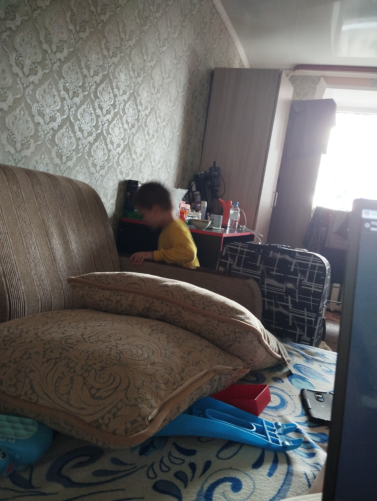 The image depicts an indoor scene, likely a living room, with a child in a yellow shirt visible near a sofa. The room is furnished with a sofa, a cabinet, and a small table with various items on it, including a water bottle and a cup. There are also two suitcases, one with a black and white geometric pattern, placed near the cabinet. The overall atmosphere appears casual and domestic. The location is identified as Kingisepp, Russia.