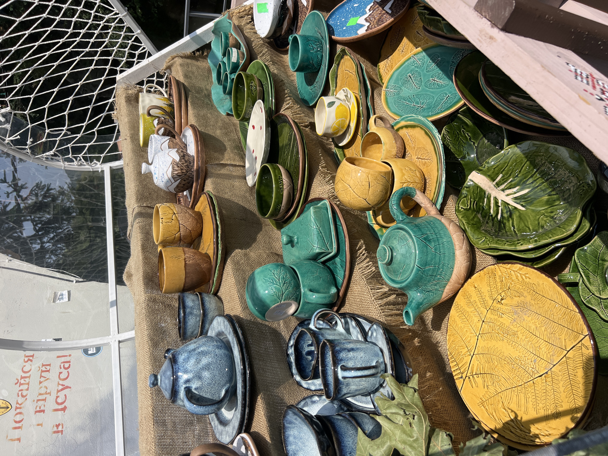 This is a close-up, eye-level outdoor shot of a display of handcrafted ceramic tableware, possibly at a market stall in Yaremche, Ukraine. The items are arranged on burlap fabric, creating a rustic presentation. The tableware includes teapots, cups, saucers, bowls, and plates. The colors are vibrant, featuring shades of blue, green, yellow, and brown, with some pieces decorated with leaf or mountain motifs. In the background, a white net structure is visible, and partially obscured text in red, likely Ukrainian, is present on a wall, reading "Покажися і вір у Ісуса!" (Show yourself and believe in Jesus!). The scene appears to be set on a sunny day, as indicated by the lighting and shadows. No people are visible in the frame.