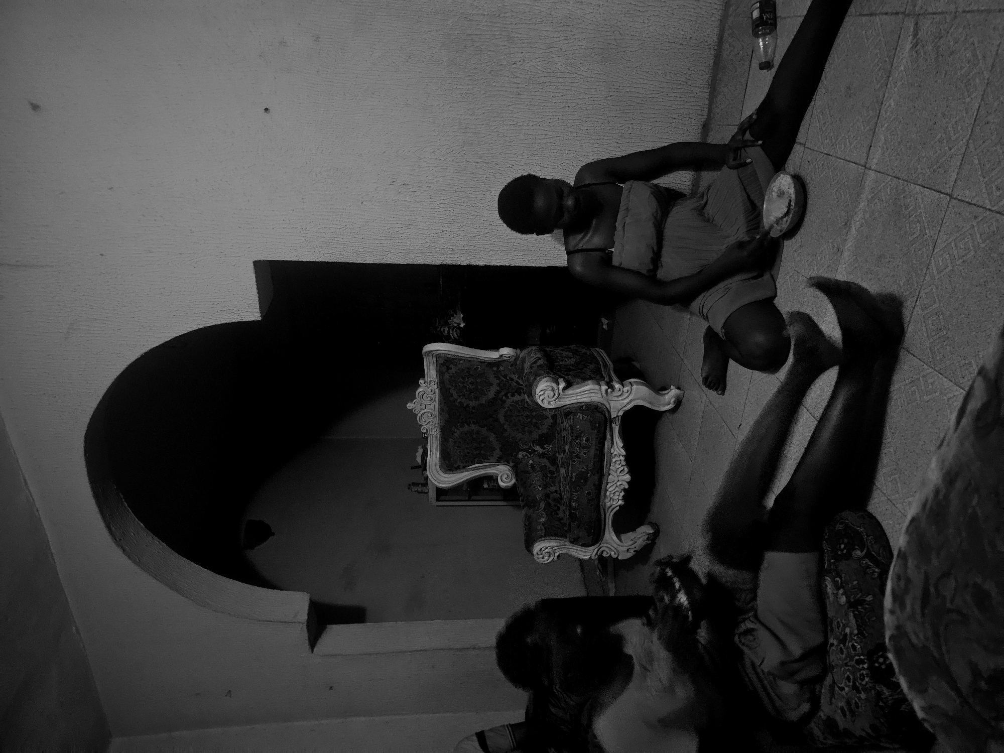 This is a monochromatic image capturing an indoor scene from an overhead, rotated perspective. In the upper right portion, a person is seated on a tiled floor, facing towards the left side of the frame. They are wearing a light-colored top and holding a light-colored bowl or dish, appearing to eat or prepare food. Their head is bowed. Near this person's extended leg, a plastic bottle is visible on the floor, with faint text, possibly "VALMIN WATER," discernible on its label.
In the bottom right, a second individual is partially visible, reclining or lying on the tiled floor with their legs extended. Their body is covered by a dark, patterned fabric, and their face is obscured.
In the mid-ground, an ornate armchair or sofa with a light-colored, possibly carved frame and dark, patterned upholstery is prominently positioned. The floor throughout the room is covered with square, patterned tiles. The walls are textured and painted. A large, dark arched doorway or opening is visible in the left background, creating a strong shadow area.
The overall lighting is dim, resulting in significant contrast and deep shadows, contributing to a subdued atmosphere. The scene suggests a casual, quiet moment, possibly at night or in a dimly lit room, with individuals relaxing or having a meal.