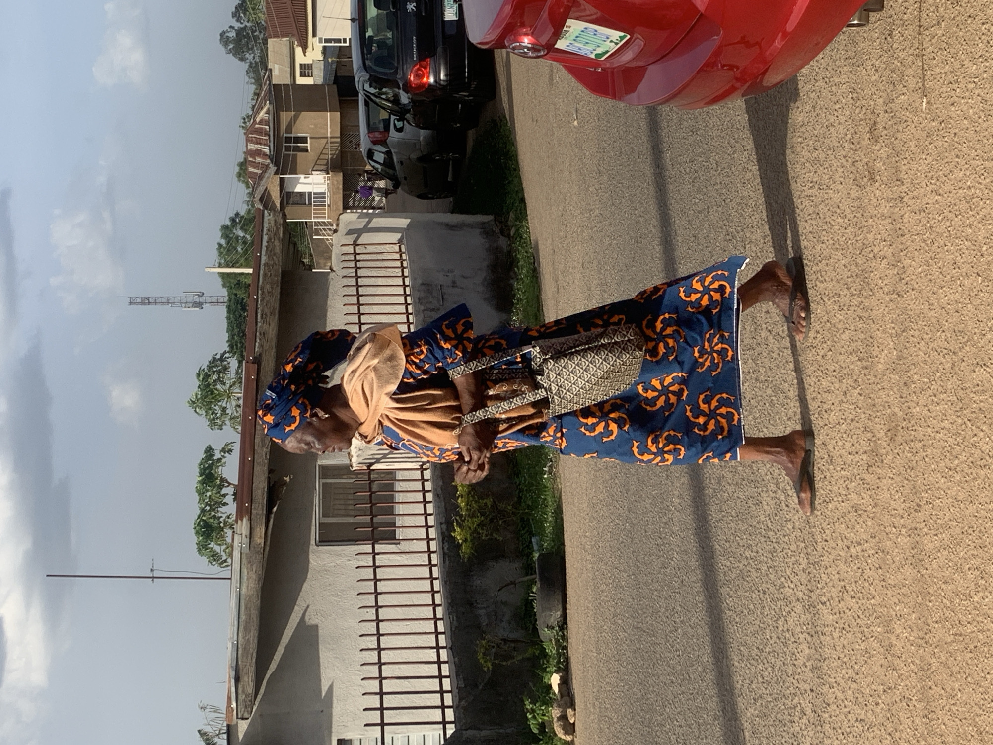An elderly woman wearing a blue and orange patterned dress and headwrap walks along a paved road in Jos, Nigeria. She carries a patterned handbag. Behind her are residential buildings with metal fences and parked cars. A communication tower is visible in the background.