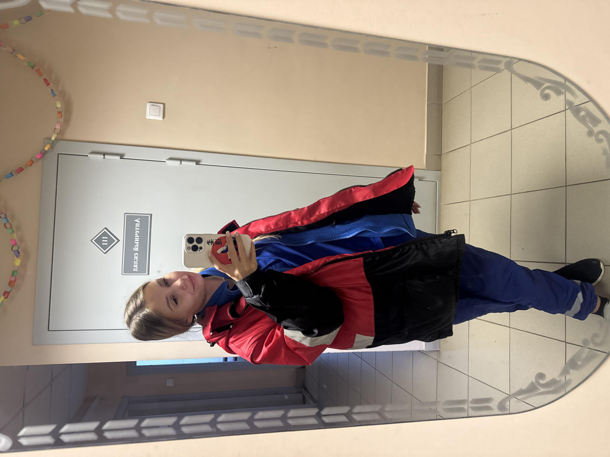A young woman is reflected in a mirror, holding a smartphone. She is wearing a blue uniform and a red and black jacket. The background features a door with Russian text, a light switch, and a chain of paper rings. The mirror is located in the city of Chaikovsky, Russia.