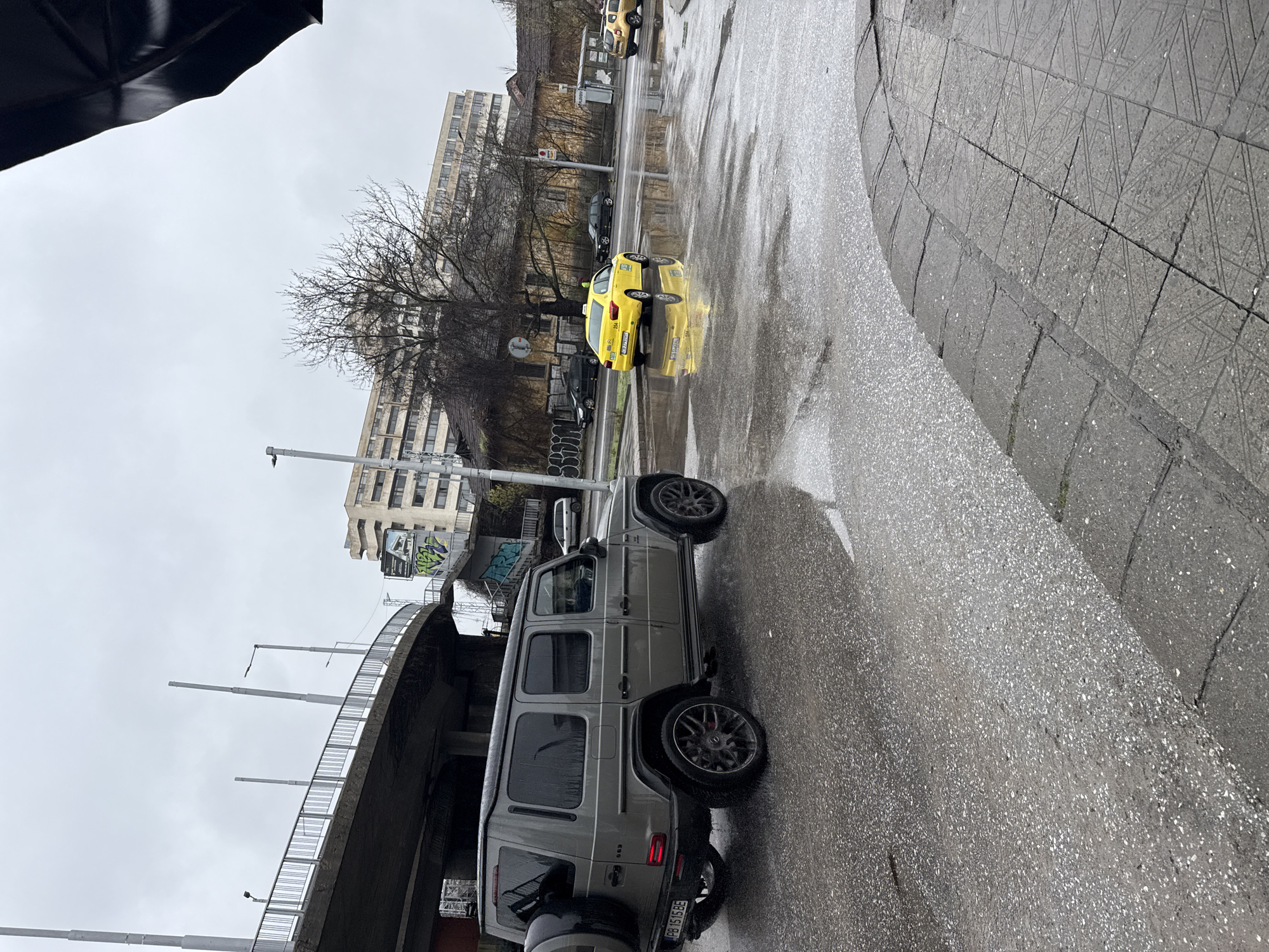 The image captures a rainy day in Plovdiv, Bulgaria. The foreground features a grey Mercedes-Benz G-Class SUV parked on a wet asphalt road beside a raised concrete bridge. A yellow taxi cab is visible in the background, reflecting in the wet pavement, and several other cars are parked further up the road. Buildings line the street, with bare trees adding to the urban landscape. The sky is overcast, and part of a black umbrella is visible in the upper-left corner, suggesting the photographer is taking cover from the rain. The sidewalk to the right is paved with patterned concrete slabs.