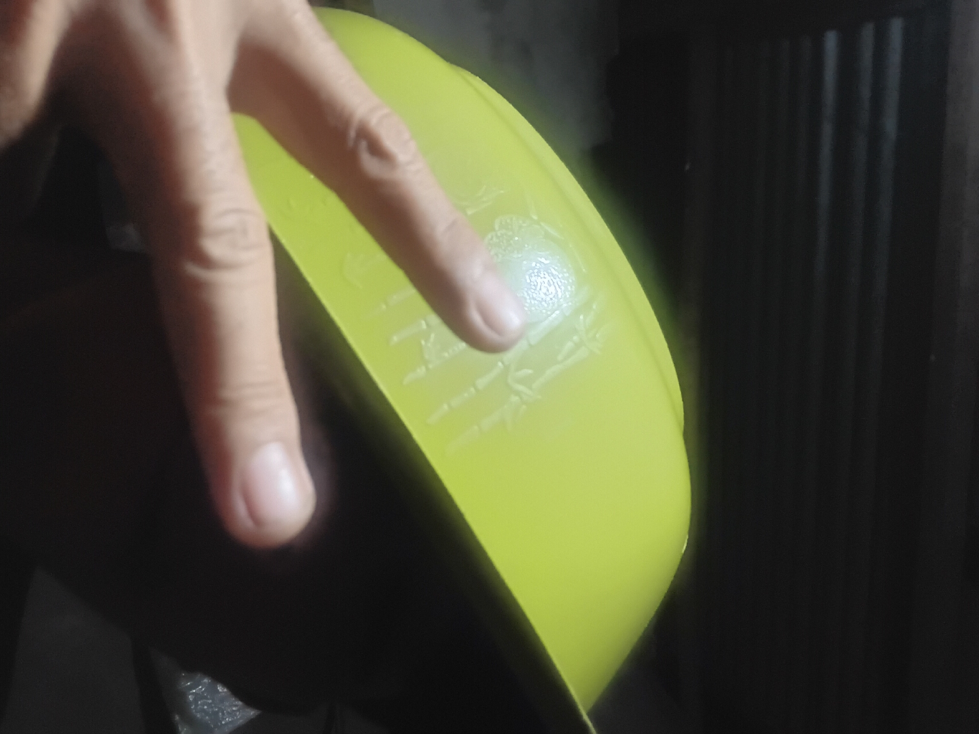 The image shows a close-up of a person's hand, with two fingers extended in a peace sign, partially obscuring a bright green bowl or basin. The bowl has an embossed design on its surface, possibly depicting bamboo stalks. The background is dark and out of focus, with horizontal lines that suggest a slatted ceiling or blinds. The lighting is dim and appears to be artificial. There is no discernible information about the location in Prabumulih, Indonesia, within the image itself.