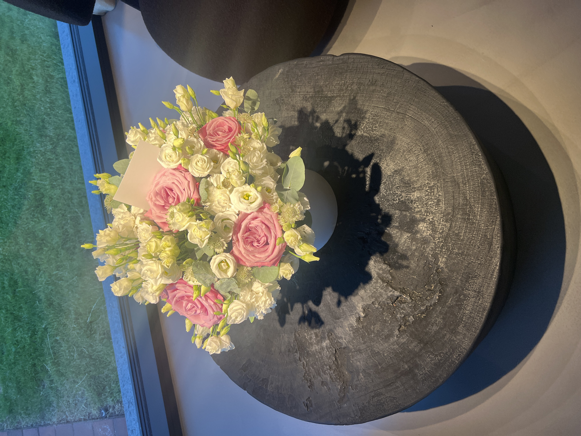 A bouquet of pink and white flowers is arranged in a white vase on a dark, circular wooden table. The table has a rough, textured surface with visible wood grain. A small, cream-colored card is placed within the flowers. The table is situated indoors, near a large window. Through the window, a patch of green grass is visible, suggesting an outdoor area beyond. The lighting in the image casts a distinct shadow of the bouquet and table onto the floor. The background is dimly lit and includes a dark, upholstered chair. The scene appears to be in a residential or commercial setting.