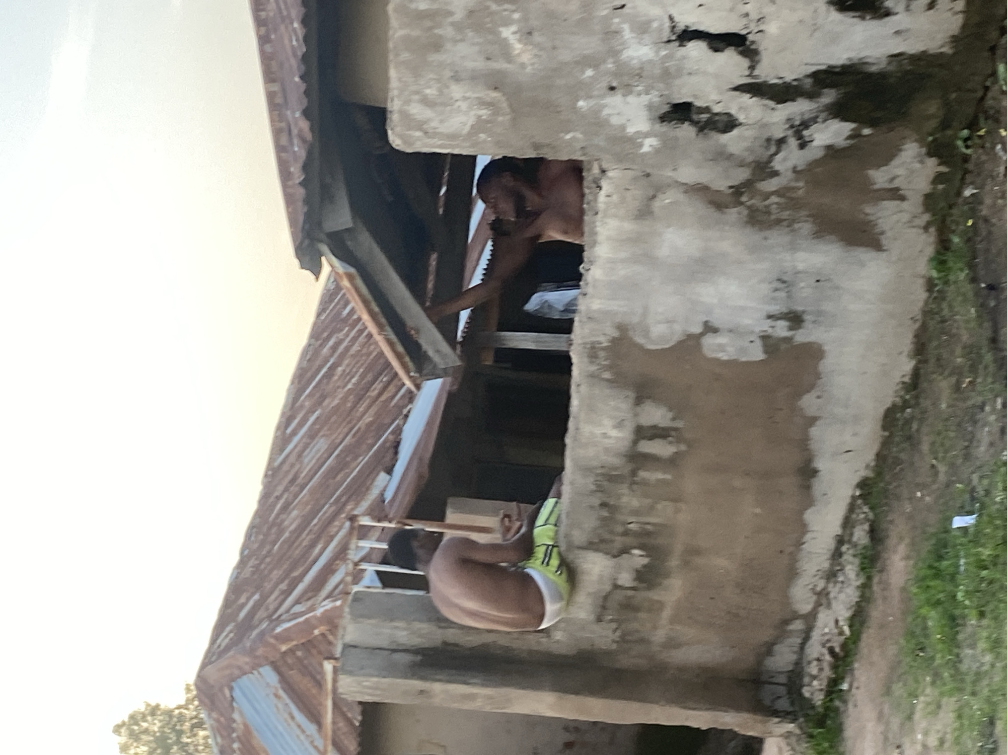 In Jauro Yinu, Nigeria, two shirtless young men are visible. One is seated on a weathered concrete wall, wearing lime green and white patterned shorts. He is facing away from the viewer and appears to be holding a thin, light-colored object vertically. The other man is reaching up towards the underside of a corrugated metal roof extension. He is partially obscured by a concrete pillar and is looking upwards. The structures appear aged, with rusty metal roofing and chipped concrete walls. The background shows a hazy sky and some greenery.