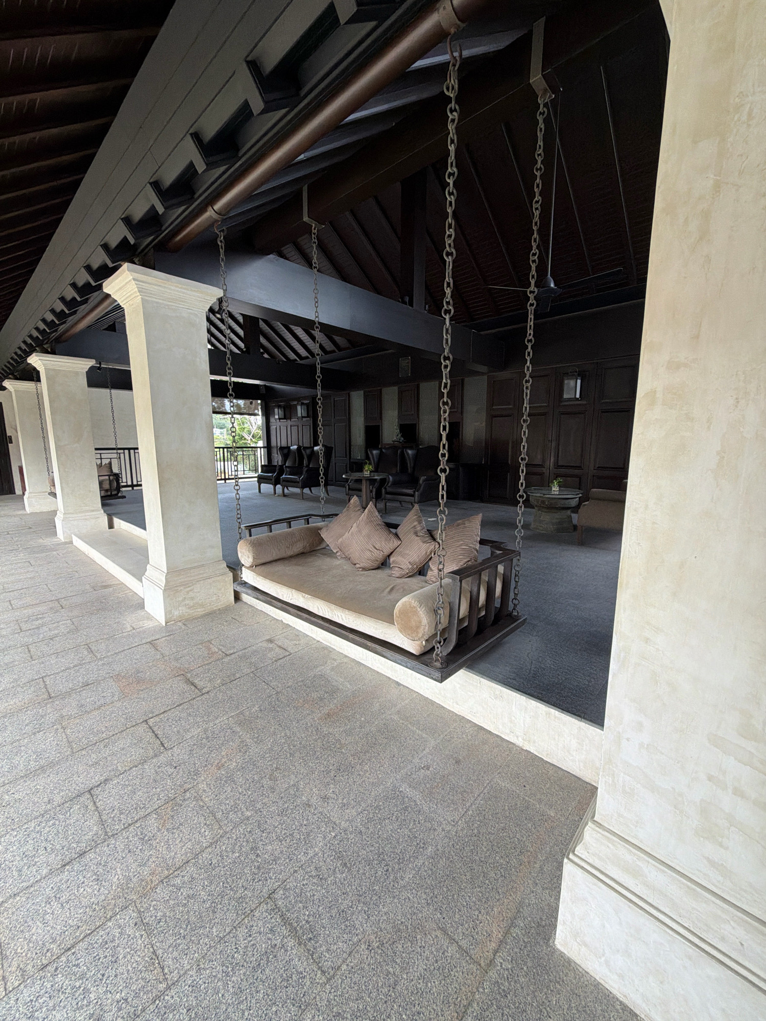 The image shows a covered outdoor seating area at what appears to be a resort in the Koh Samui District of Thailand. In the foreground, a cushioned daybed swing hangs from metal chains, supported by a dark wooden beam. The swing is upholstered in a light brown fabric and has several matching cushions. The floor is paved with large, grey granite tiles. To the left, there are large, white, square pillars. Behind the swing, the area is furnished with dark wooden paneling on the walls, several dark leather armchairs, and a round stone table with a small plant in the center. Natural light illuminates the scene, suggesting it is daytime. In the background, through an open doorway, a glimpse of greenery and a railing is visible.
