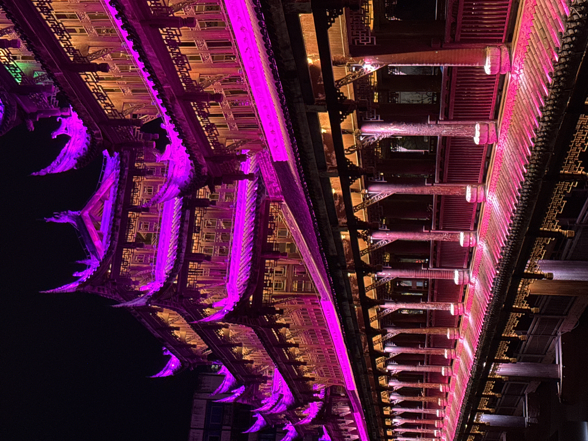 This is a low-angle, color photograph of an illuminated traditional Chinese architectural structure at night. The location is likely in Qianjiang, China, as suggested by the architectural style. The building is ornate, with dark wooden beams and intricate latticework. Portions of the structure are lit with vibrant pink-purple light, creating a stark contrast against the dark night sky. Some areas are lit with a warmer, yellowish light, adding depth to the scene. The roofline has upturned eaves, typical of Chinese architecture. There are no people visible in the scene. The overall impression is one of a historic building modernized with artistic lighting.