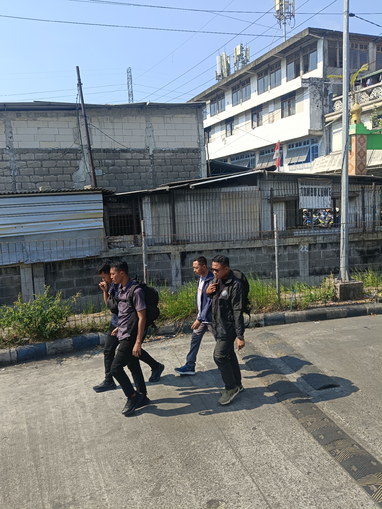 In Kedungrejo, Indonesia, four men are walking across a paved road during daylight. The men are positioned from left to right as they move away from the viewer. The first man, on the far left, is wearing a dark blue patterned shirt and black pants. The second man, slightly ahead and to the right, wears a dark purple collared shirt with short sleeves and black pants. He is carrying a black backpack. The third man, to the right of the second, is dressed in a blue and white jacket over a light blue shirt and gray pants. He also carries a black backpack. The fourth man, on the far right, is wearing a black jacket and black pants, and is carrying a black backpack. They are walking on a road with speed bumps. In the background, there are various structures including a multi-story building with multiple windows, a smaller building with corrugated metal siding, and a brick wall. Vegetation, including tall grass and bushes, is visible along the side of the road behind a fence. The sky is clear and blue.