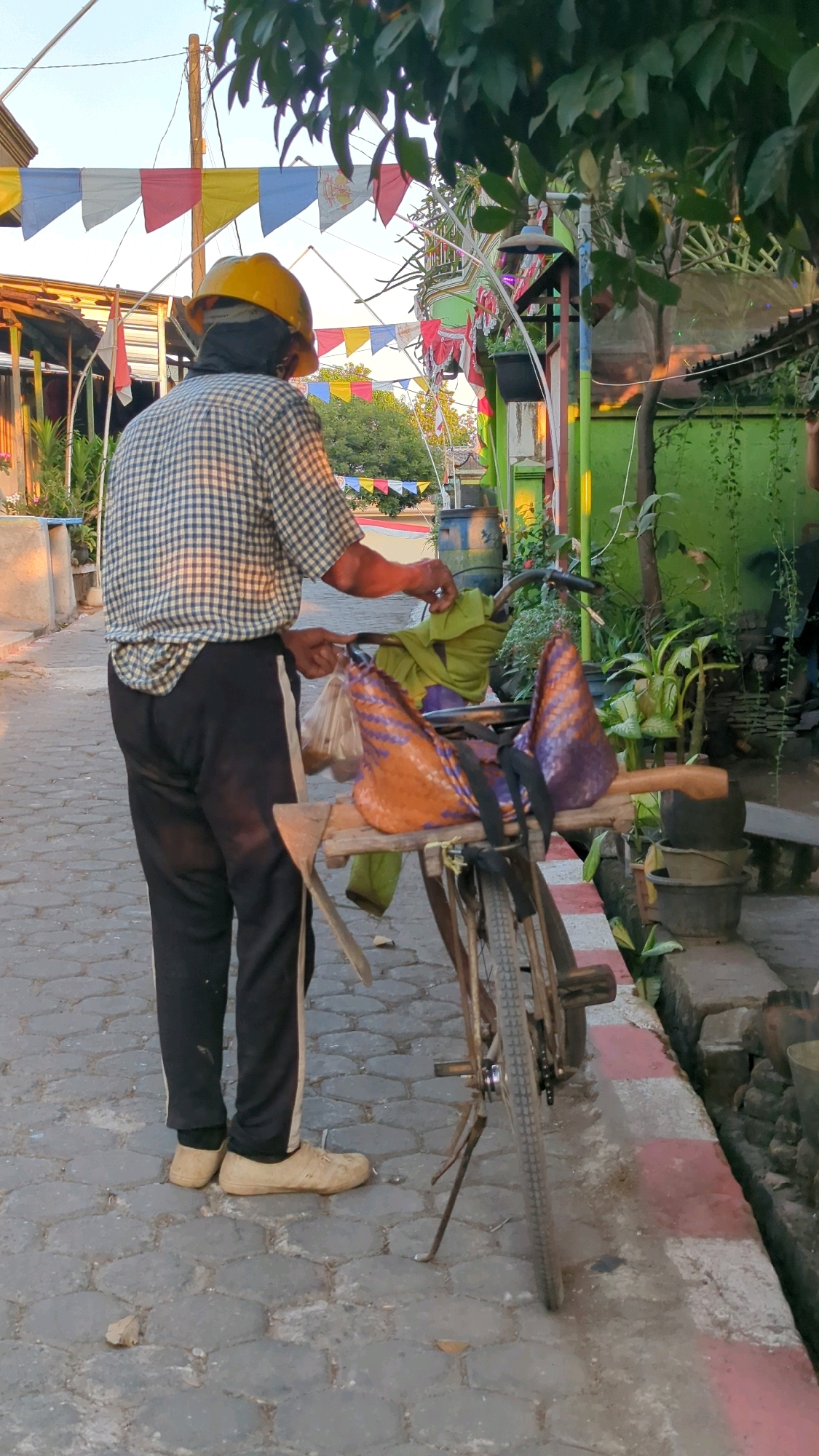 A lone individual, wearing a yellow hard hat, a blue and white checkered shirt, and black pants with a white stripe, stands with their back to the camera, holding a plastic bag in their right hand and interacting with a bicycle. The bicycle is laden with two woven conical bags, one orange and the other purple, secured to a wooden platform attached to the rear of the bike. A green cloth is draped over the handlebars. The scene appears to be outdoors, on a paved pathway lined with red and white curbs. In the background, colorful triangular flags are strung across a wire, and various buildings, plants, and a large blue barrel are visible. The lighting suggests it is daytime, possibly late afternoon, with some shadows present. The overall setting resembles a street or pathway in Proto, Indonesia, characterized by the vibrant flags, local architecture, and the presence of a bicycle used for transport or commerce.