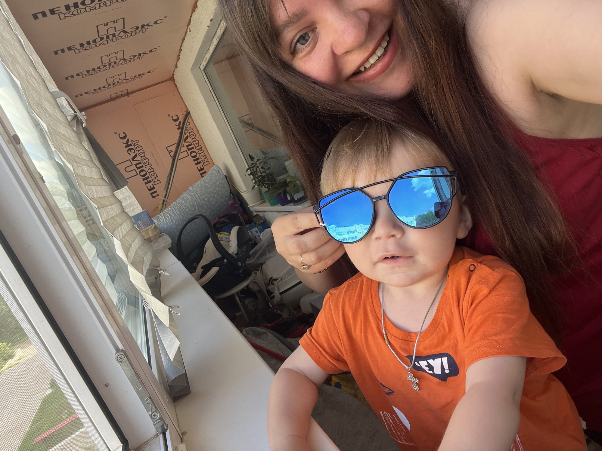 A woman and a child are taking a selfie on a balcony in Noviy, Russia. The woman, in the background, has long brown hair and is wearing a red top. She is smiling broadly. The child, in the foreground, has blond hair and is wearing blue mirrored sunglasses and an orange t-shirt with the word "HEY!" printed on it. A silver necklace with a cross pendant is visible around the child's neck. The balcony is enclosed with windows and appears to be under construction, with insulation material visible on the ceiling and walls. In the background, on the floor, are various objects including bags, shoes, and what appears to be a baby car seat. The view from the balcony shows greenery and a building.