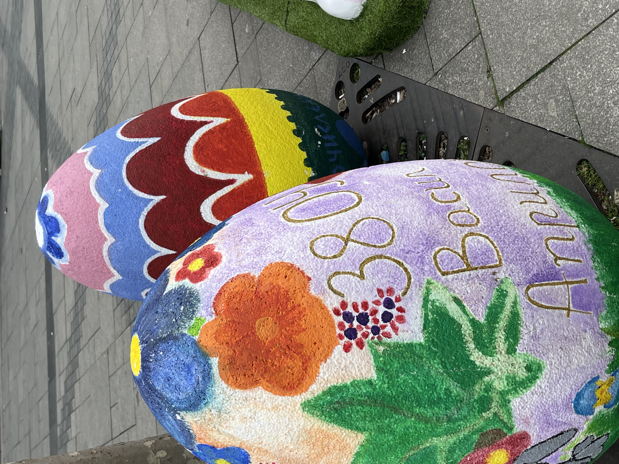 Two large, painted Easter eggs rest on or near a metal bench in an outdoor setting. The first egg features colorful patterns, including pink, blue, red, yellow, and green sections adorned with white scallops. The second egg is lavender with floral designs and the words "Beaucu 30 Ocul". There's a small patch of green grass visible in the background, and the ground is paved with grey tiles. The location is the city of Sofia, Bulgaria.