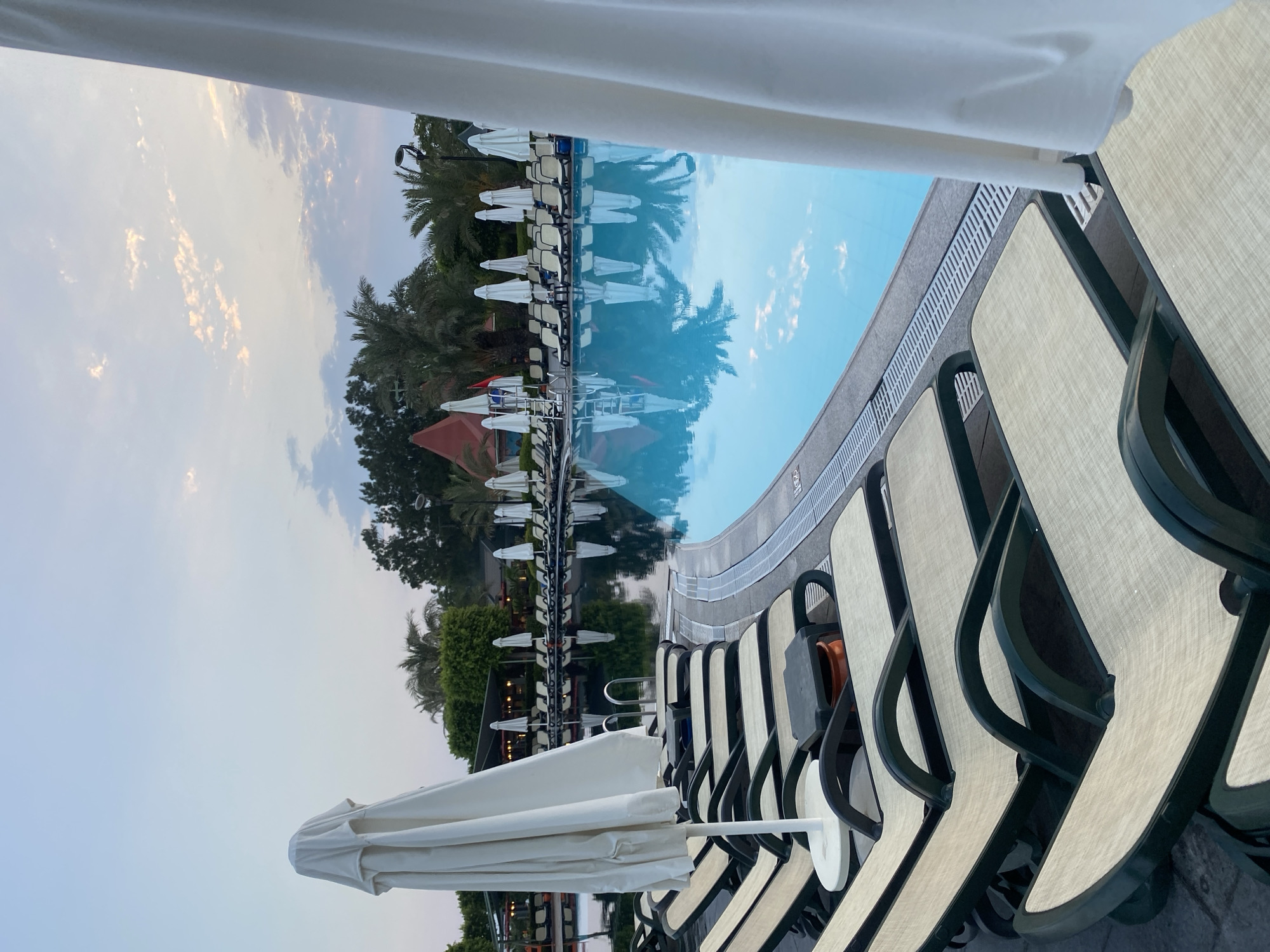 An outdoor swimming pool area is depicted at dusk, likely in Aksu, Turkey, given the context. The foreground is dominated by a row of empty, reclined lounge chairs with beige mesh seats and dark green frames, neatly arranged along the poolside. Several closed white umbrellas are positioned behind the lounge chairs. The swimming pool itself is a prominent feature, with its bright blue water reflecting the sky and surrounding palm trees and foliage. The water's surface is calm, with clear reflections visible. The edge of the pool is lined with grey paving stones and a grated drainage system. In the background, more lounge chairs and umbrellas are visible, along with a cluster of trees, including palm trees, and some structures with reddish-brown conical roofs. The sky above is a pale blue, transitioning to a softer hue near the horizon, with scattered wispy clouds that are tinged with pink and orange from the setting sun. The lighting suggests it is either sunrise or sunset. There are no people visible in the image.