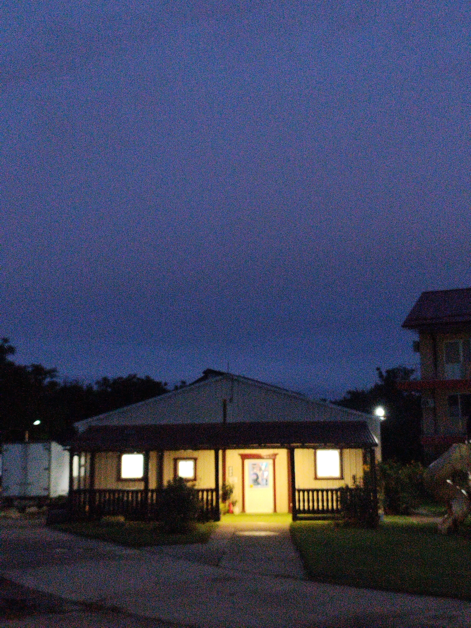 The image depicts a dimly lit scene at dusk or night, with a building that appears to be a house or a small office. The building has a covered porch with railings and two illuminated windows. The sky is a deep blue-purple, indicating low light conditions. Trees are visible in the background, silhouetted against the sky. To the right, another building is partially visible, suggesting a residential or commercial area. The ground in front of the main building is paved with what looks like concrete, and there is a patch of grass. The overall impression is one of quietness, possibly an establishment that has closed for the day or a residential property. There are no people visible in the image.