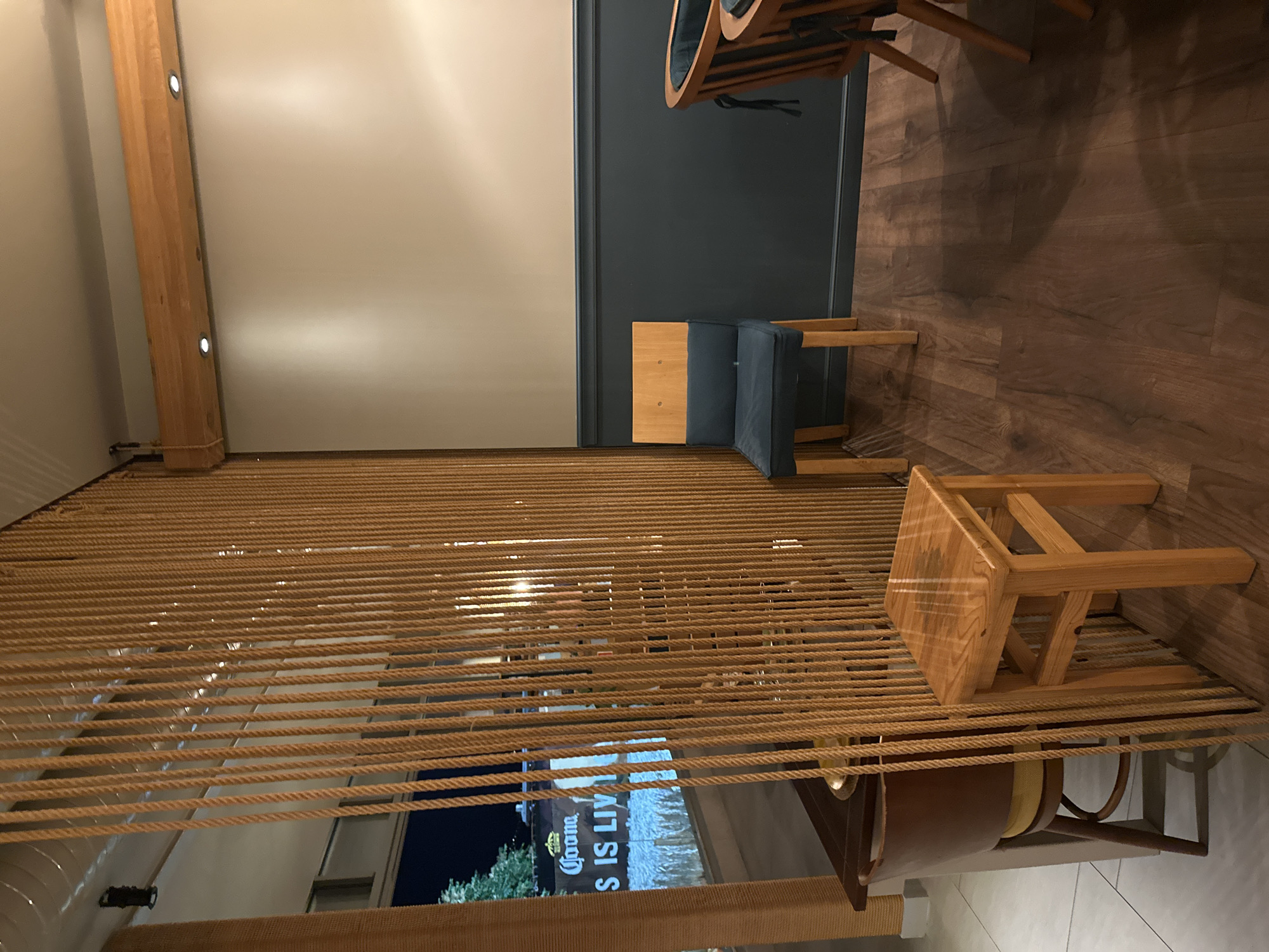 The image shows the interior of a restaurant or bar with a partition made of vertical ropes. In the foreground, there is a small wooden stool. Behind the stool and to the left, a table with a dark surface and a yellow cushion on a chair is partially visible. To the right of the rope partition, a wooden chair with a dark blue cushion is positioned against a dark gray wall. Further to the right, the back of another wooden chair with cushions is visible. The floor is made of dark wood planks. In the background, through an opening in the rope partition, a sign with "Corona S IS LIV" is illuminated, suggesting an outdoor or semi-outdoor area. The overall lighting is dim, with spotlights visible on a wooden beam above the wall. There are no people visible in the image.