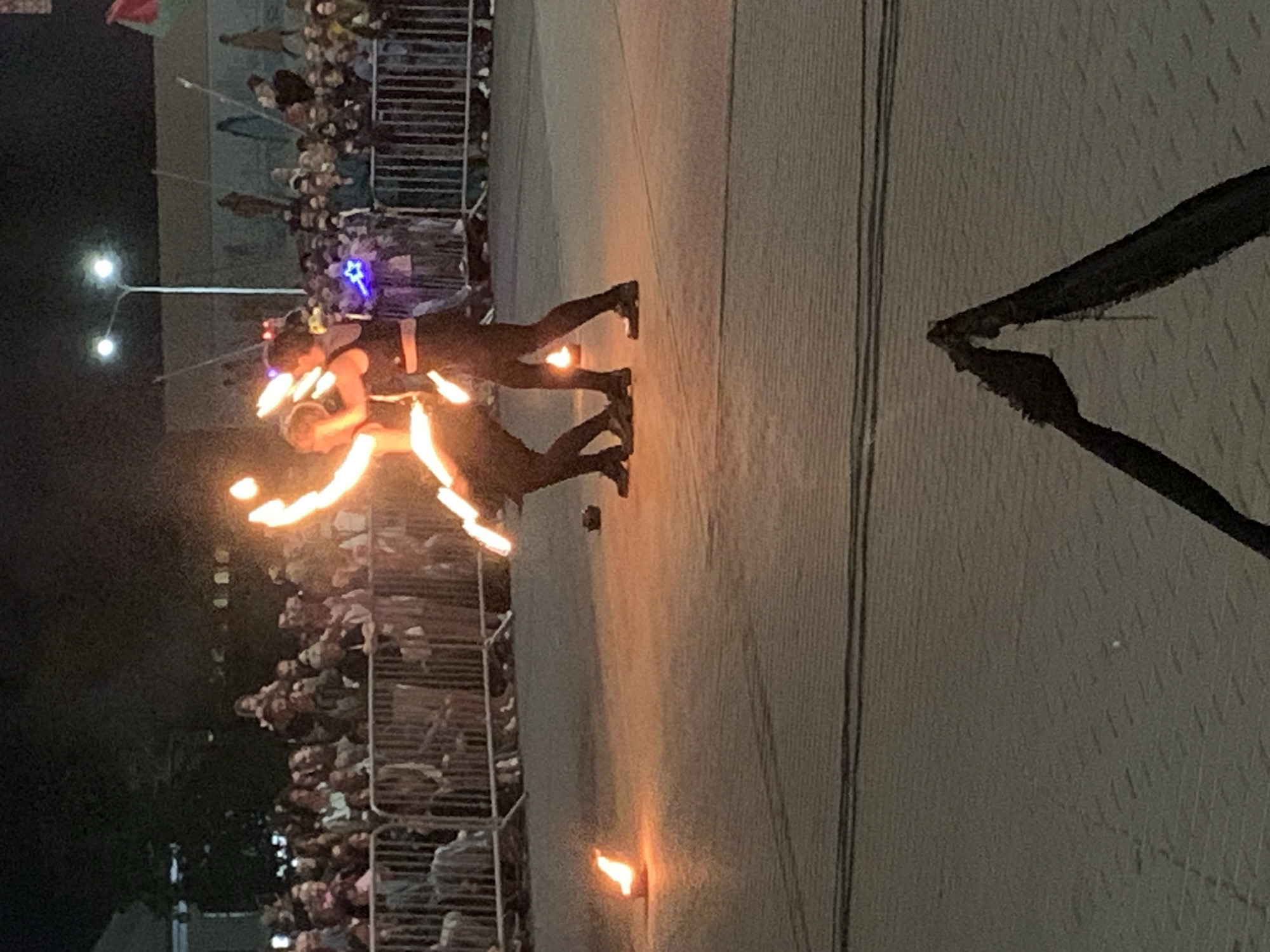 This image captures a nighttime fire performance in the city of Kapyĺ, Belarus, likely in a public square or open area. Two performers, dressed in dark clothing, are actively engaged in spinning flaming props, creating bright arcs of fire in the darkness. One performer, on the right, appears to be a male in dark pants and a black tank top. The other, on the left, is in a similar dark outfit. In the background, a crowd of spectators is visible behind a metal barrier, suggesting a public event or festival. Streetlights and the glow from the fires illuminate the scene. The ground is paved, and in the foreground, a dark, triangular object, possibly part of a stage prop or equipment, is visible.
