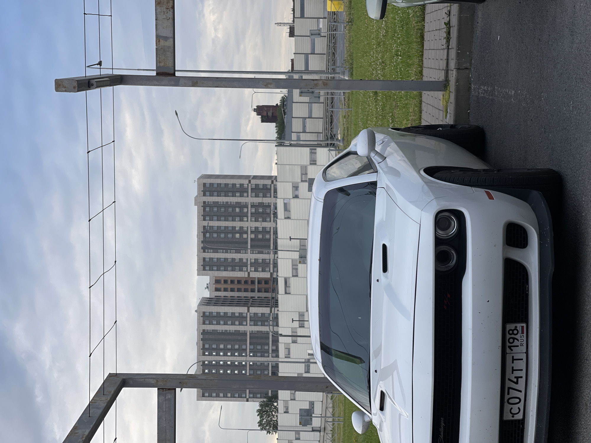 A white Dodge Challenger is parked in a parking lot. The car is facing forward, with its front bumper and license plate visible. The license plate number is C074TT 198. In the background, there are tall apartment buildings and a highway sound barrier. The sky is cloudy.