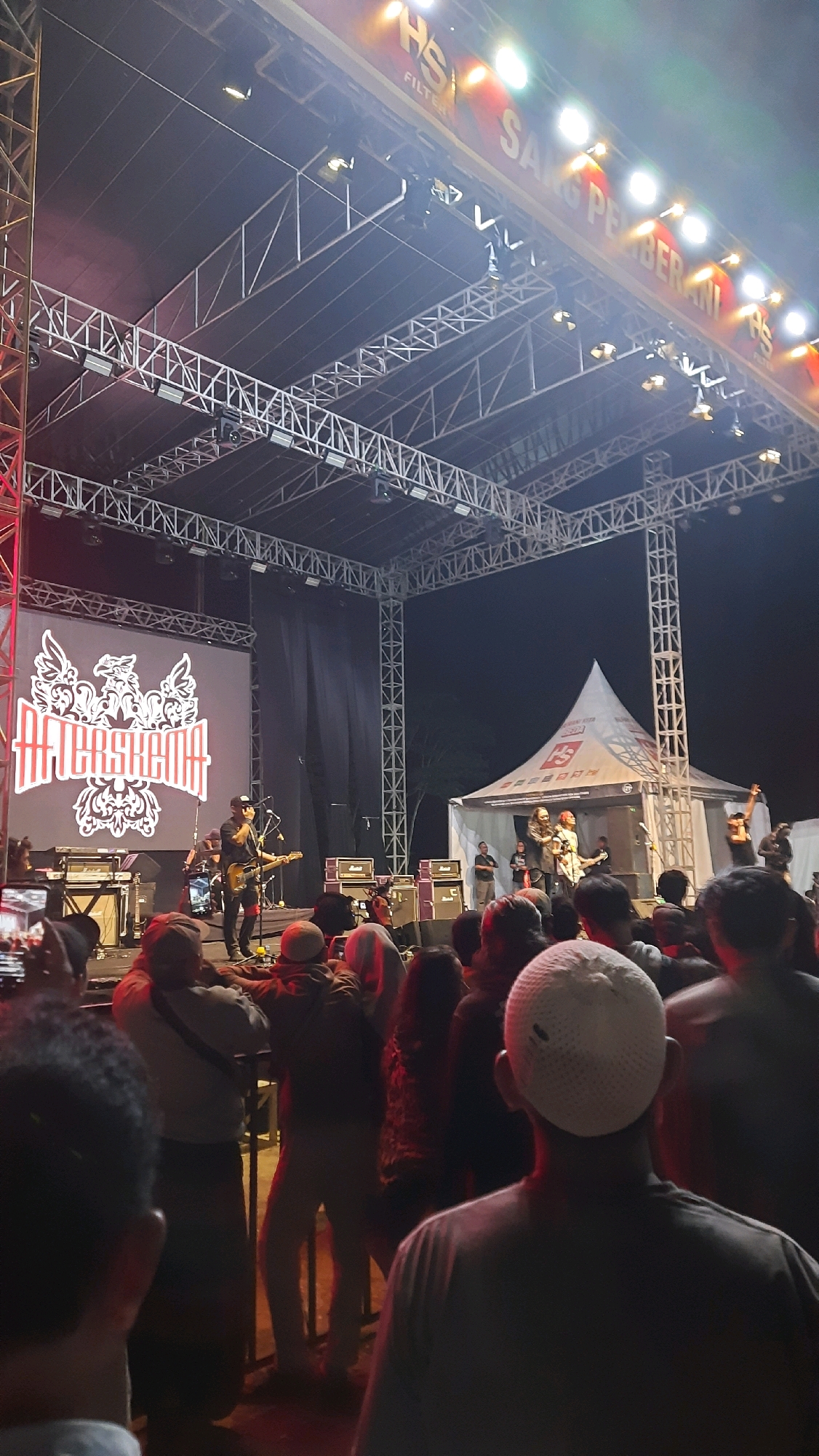 This image captures an outdoor concert event at night in Minggir, Indonesia. A band, identified by the logo "AFTERSHOCK" on a large screen, performs on a stage. The stage is constructed with metal scaffolding and illuminated by stage lights. Several band members are visible, including a guitarist in the foreground and others on stage. Amplifiers and other musical equipment are arranged on the stage. In front of the stage, a crowd of spectators is gathered, many of whom are facing the stage and holding up phones, presumably to record or take photos. To the right of the stage, a white tent with logos is visible. The atmosphere appears energetic, with people enjoying the live music. The overall scene suggests a lively community event.