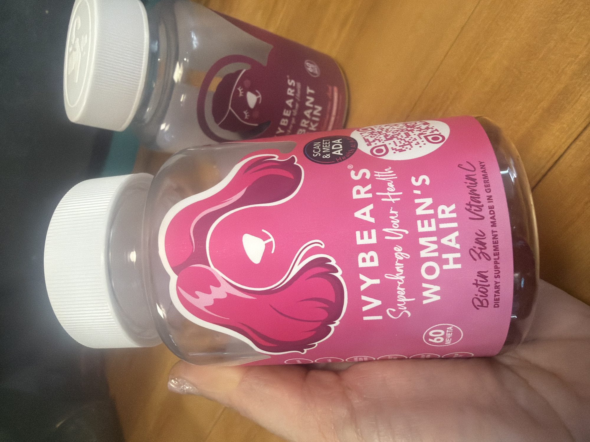 The image shows two bottles of IVYBEARS dietary supplements. The bottle in the foreground is labeled "Women's Hair" and contains biotin, zinc, and vitamin C. The label has a QR code and reads "Supercharge Your Health". The bottle in the background is labeled "YBEARS Brant Kin". The bottles have white lids and are clear, revealing the gummy bears inside. A person's hand with a glittery nail is holding the front bottle. The background is blurry with some brown and black objects visible. The bottles are lying on a wooden surface. Sofia, Bulgaria, is not depicted in this image.