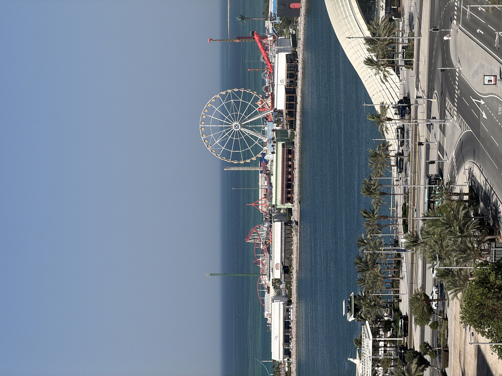 This is an eye-level, medium shot of an amusement park situated on a waterfront. The park features a large Ferris wheel, roller coasters, and other rides, all set against a backdrop of the ocean and a clear blue sky. In the foreground, there is a paved area with palm trees, a street with cars and a bus, and a curved white building. The scene appears to be a sunny day in an urban coastal area.