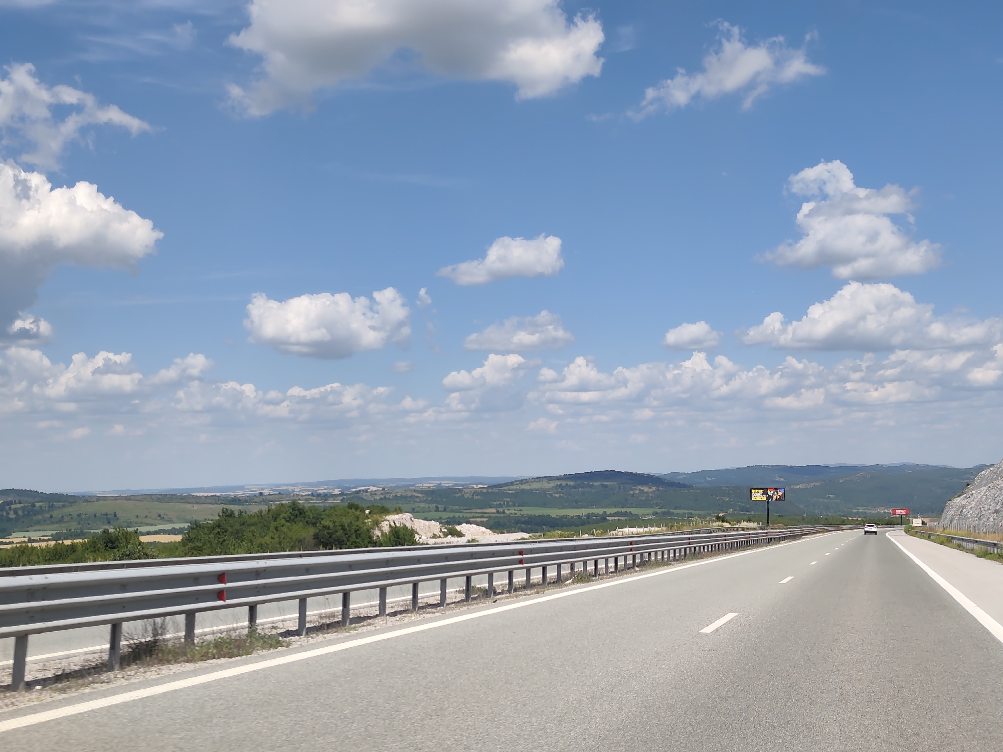 The image depicts a highway scene in a rural, mountainous landscape under a bright, partly cloudy sky, suggesting daytime. The road, paved with asphalt and marked with white lines, curves gently to the right and stretches into the distance. A metal guardrail runs along the left side of the road, separating it from a vegetated slope with scattered rocks. Beyond the slope, a rolling landscape unfolds with fields, trees, and distant hills or mountains.
In the far distance, a white car is visible on the highway, indicating that the photo was taken from another vehicle traveling on the same road. There are two billboards visible along the side of the road. The billboard on the left appears to be an advertisement with images of people, and the billboard on the right is red with some white text that is too blurry to read. The overall scene conveys a sense of travel through a scenic, undeveloped area. The weather appears to be fair and sunny, with fluffy white clouds dotting the blue sky. The terrain suggests a location in Bulgaria, specifically Yablanitsa, given the provided context. No people are directly visible in the image.