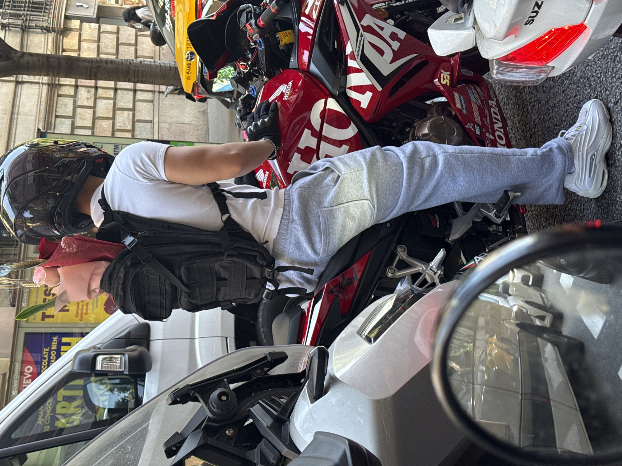 The image shows a person sitting on a red and white Honda motorcycle in Barcelona, Spain. The person is wearing a black helmet, a white t-shirt, gray sweatpants, and white sneakers. They have a black backpack and gloves. A bouquet of flowers is visible. The motorcycle is parked in a busy area with other vehicles, including a white van and another motorcycle. Advertisements are visible on buildings in the background. The image is taken from a close perspective, with part of another motorcycle visible in the foreground.