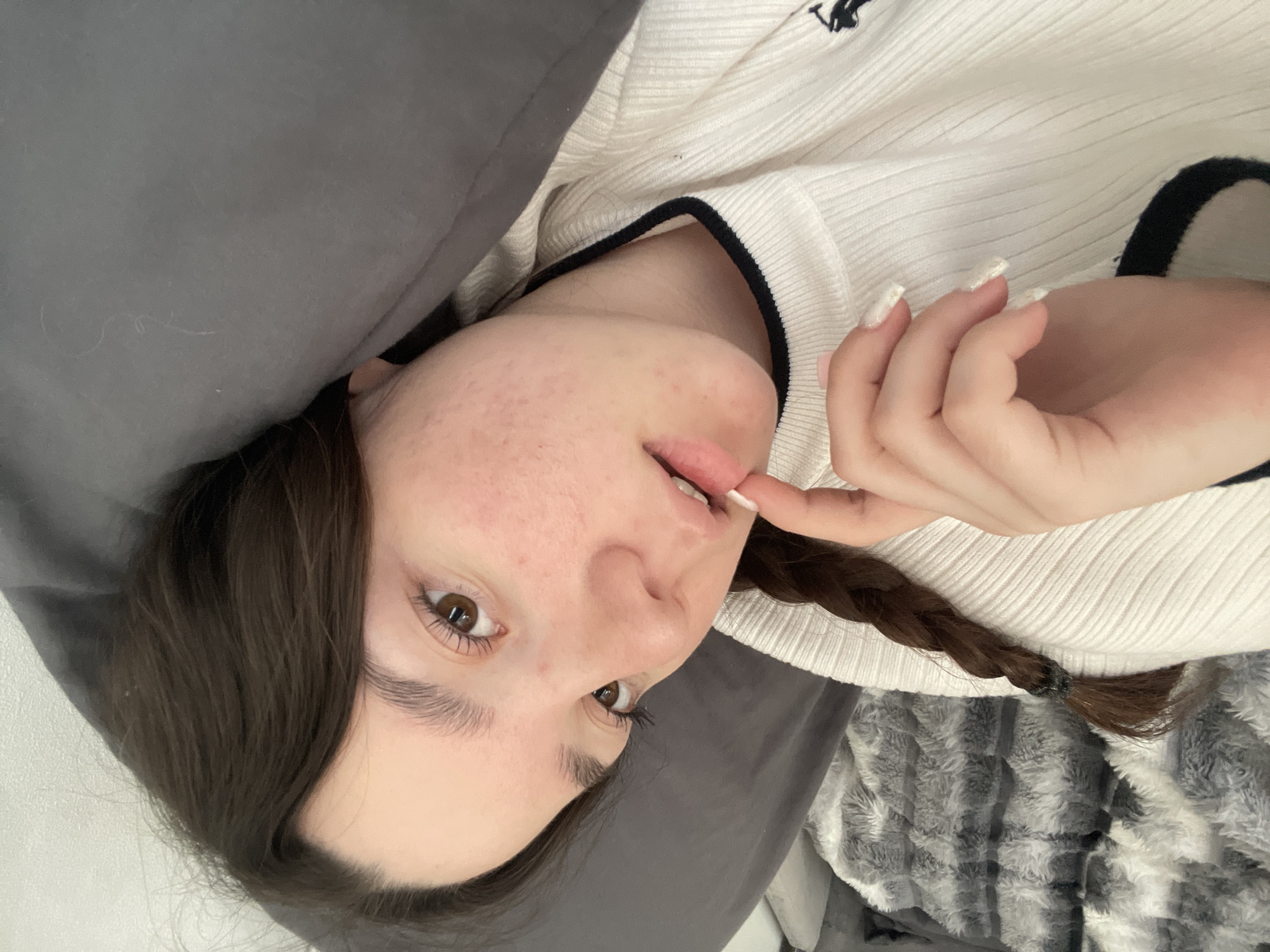 A close-up, rotated selfie of a young woman with light skin, brown eyes, and long brown hair partially braided. She is lying down with her head on a gray pillow and draped with a gray and white blanket. She is wearing a white sweater with black trim and has long, artificial nails. Her right index finger is touching her lower lip. The background includes a partial view of a white wall. The location of Chaikovsky, Russia, is not discernible from the image.
