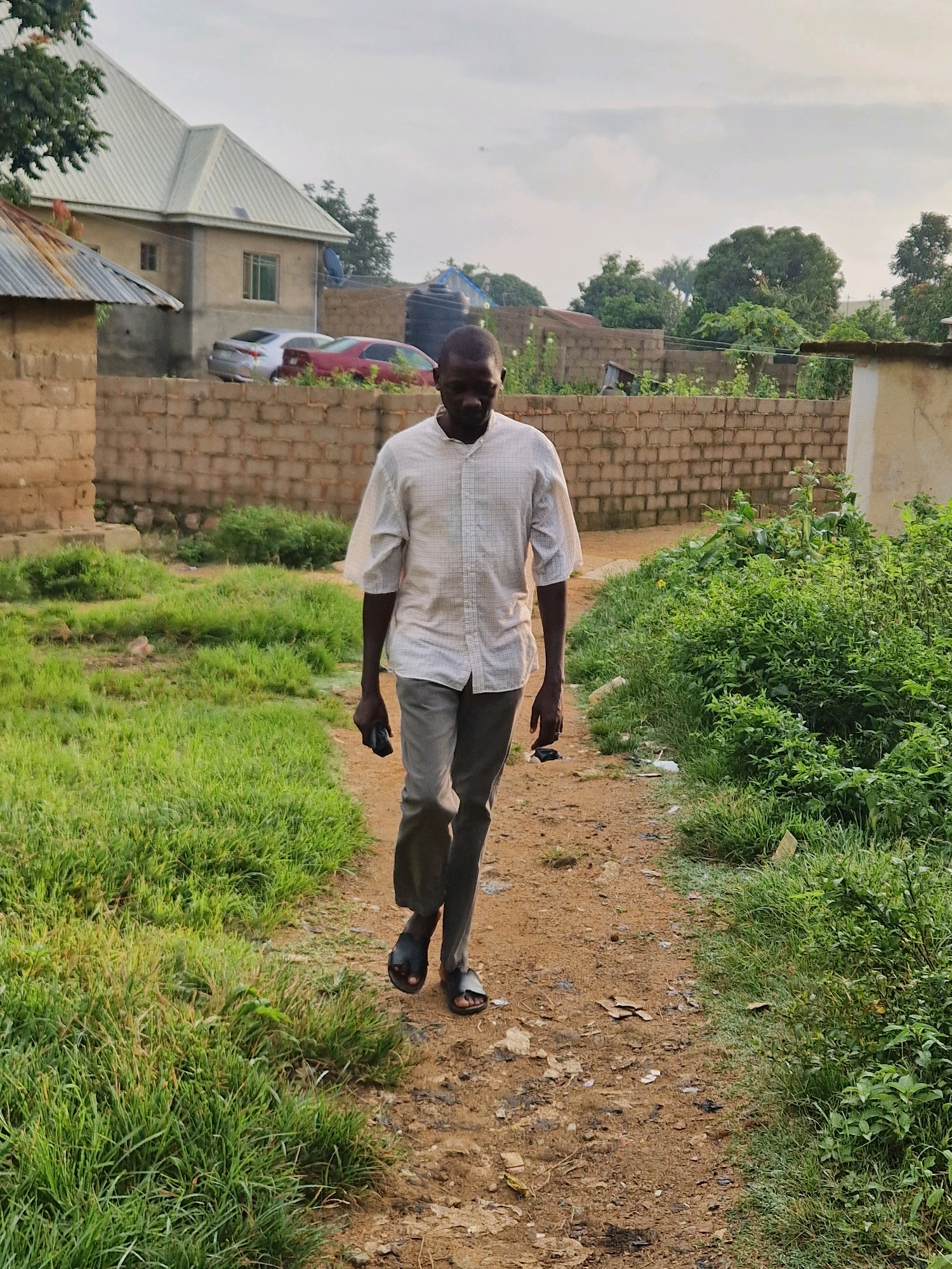 A man walks down a dirt path in a residential area in Jos, Nigeria. The man is wearing a short-sleeved collared shirt and light-colored pants. He is holding a dark-colored object, possibly a phone, in his right hand. The path is lined with tall green grass on the left and a dense patch of vegetation on the right. In the background, there are brick walls, houses with corrugated metal roofs, and parked cars. The sky is overcast.