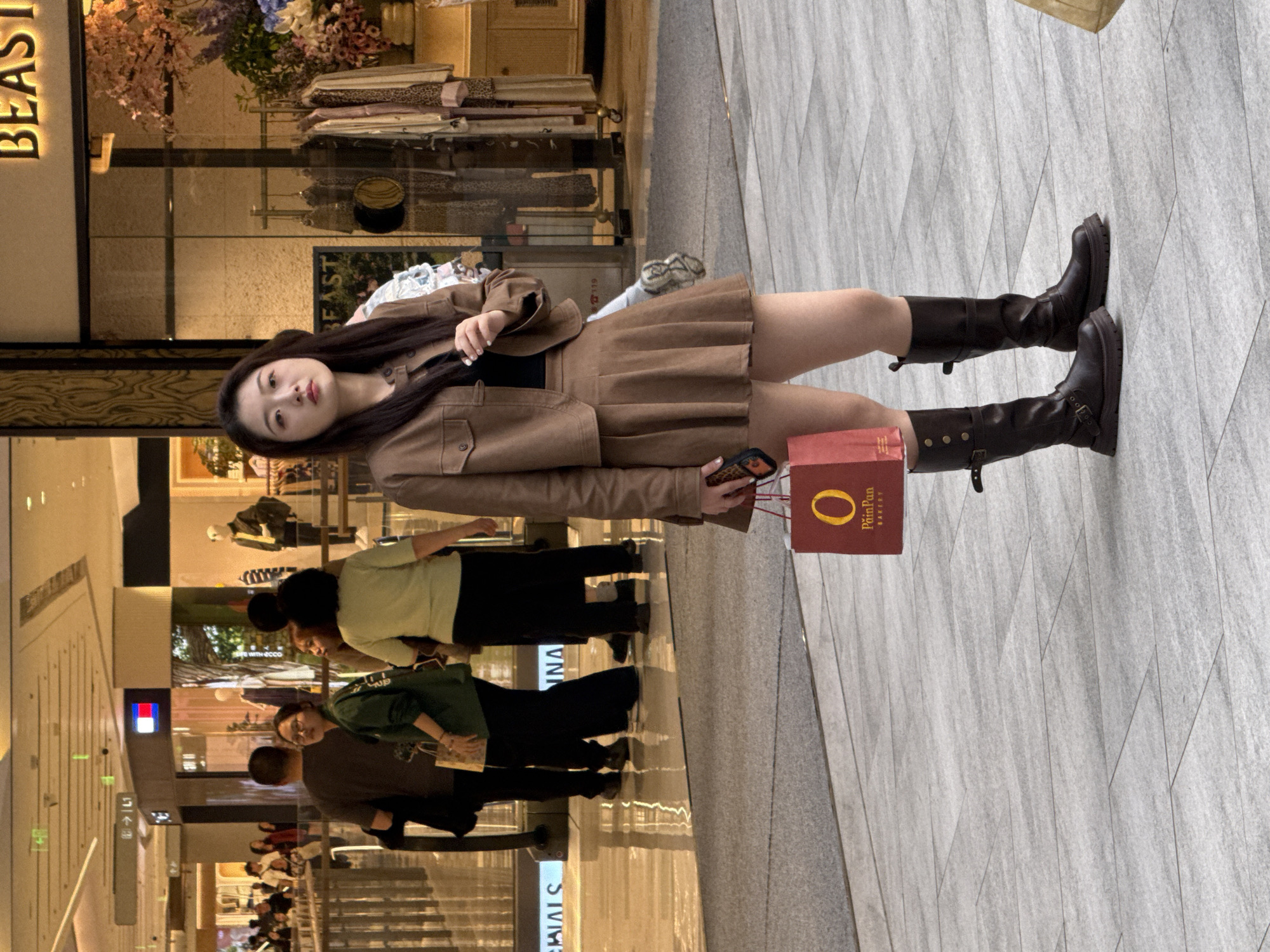 The image shows a young woman in a brown skirt suit and dark knee-high boots posing in what appears to be a shopping mall in Liangjiang, China. She is holding a red PainPan Bakery shopping bag. Several other people are walking by in the background.