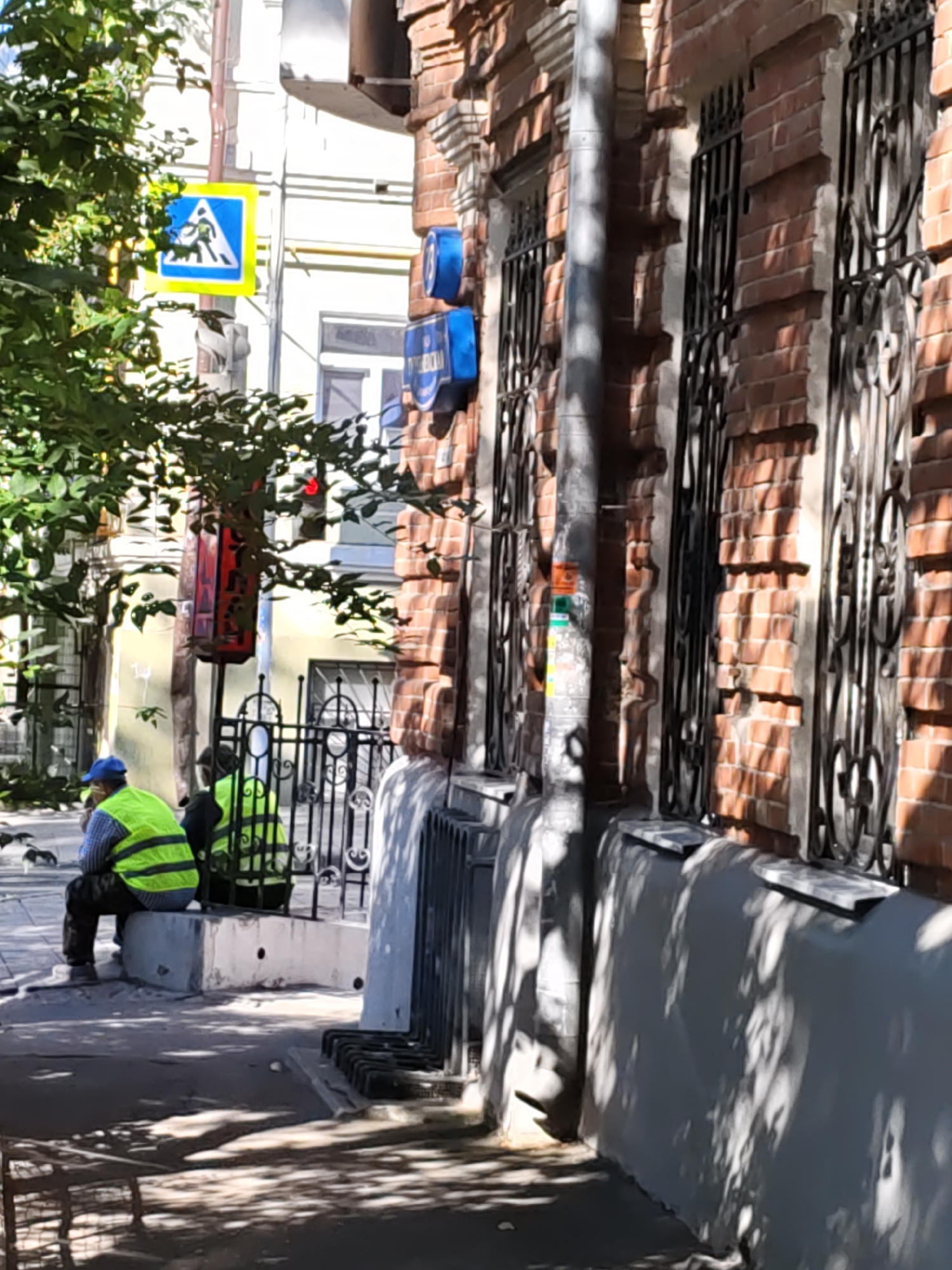 The image depicts a street scene in Rostov-on-Don, Russia, with a focus on a brick building facade. In the foreground, two individuals wearing high-visibility vests are present, one seated on a concrete ledge and the other standing nearby. Behind them, a traffic sign with a pedestrian crossing symbol is visible amidst leafy green trees. The building itself is constructed of red brick and features ornate wrought-iron window grates. A street lamp is also present, with a red light visible at its top. The ground is paved, and shadows from trees are cast across the scene, indicating a sunny day. There are also several small, dark objects on the ground, possibly birds or litter, in front of the seated individual.