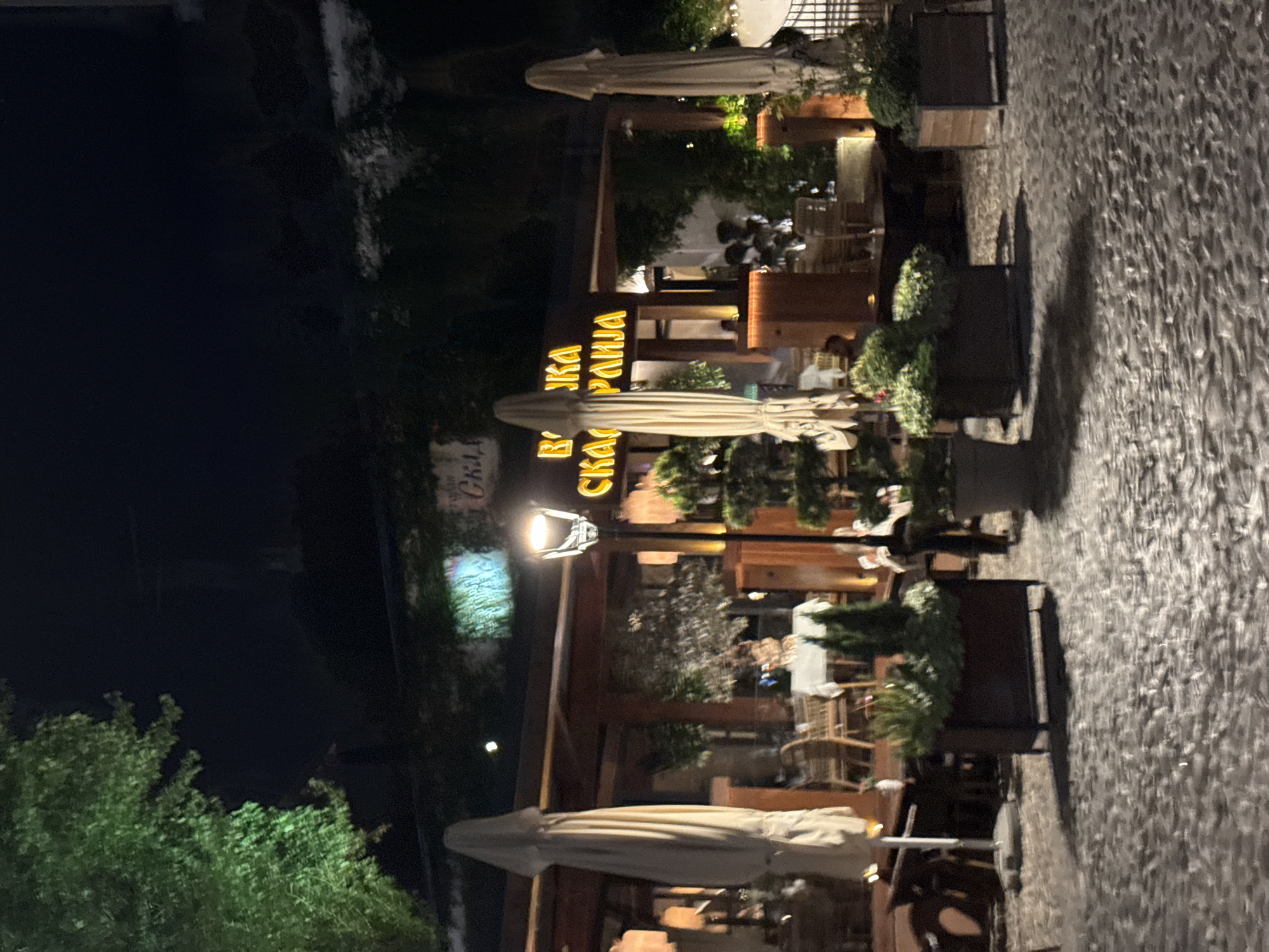 The image displays an outdoor dining area of a restaurant or cafe at night, illuminated by artificial light sources. The scene is observed from a slightly elevated perspective, looking across the patio.
In the foreground and midground, several closed, light-colored outdoor umbrellas stand upright. These are positioned among various tables and chairs, some of which are visible, suggesting an active dining establishment. Planters filled with green bushes and small trees are strategically placed around the seating area, enhancing the outdoor ambiance.
A prominent street lamp, emitting a bright light, stands in the midground, casting illumination on the surrounding paving, which appears to be cobblestone or textured stone. The background features a building facade with a brightly lit, yellow sign in Cyrillic script. When correctly oriented, the visible text on the sign clearly reads "МАКЕДОНСКА КУЋА", translating to "Macedonian House". Blurry figures of people can be seen seated at tables in the background, indicating patrons enjoying an evening out.
The time of day is clearly night, evidenced by the dark sky and the reliance on artificial lighting from the street lamp and the restaurant sign. The atmosphere appears relaxed and inviting, typical of an outdoor dining setting in an urban environment. The location context provided indicates this scene is in Belgrade, Serbia.